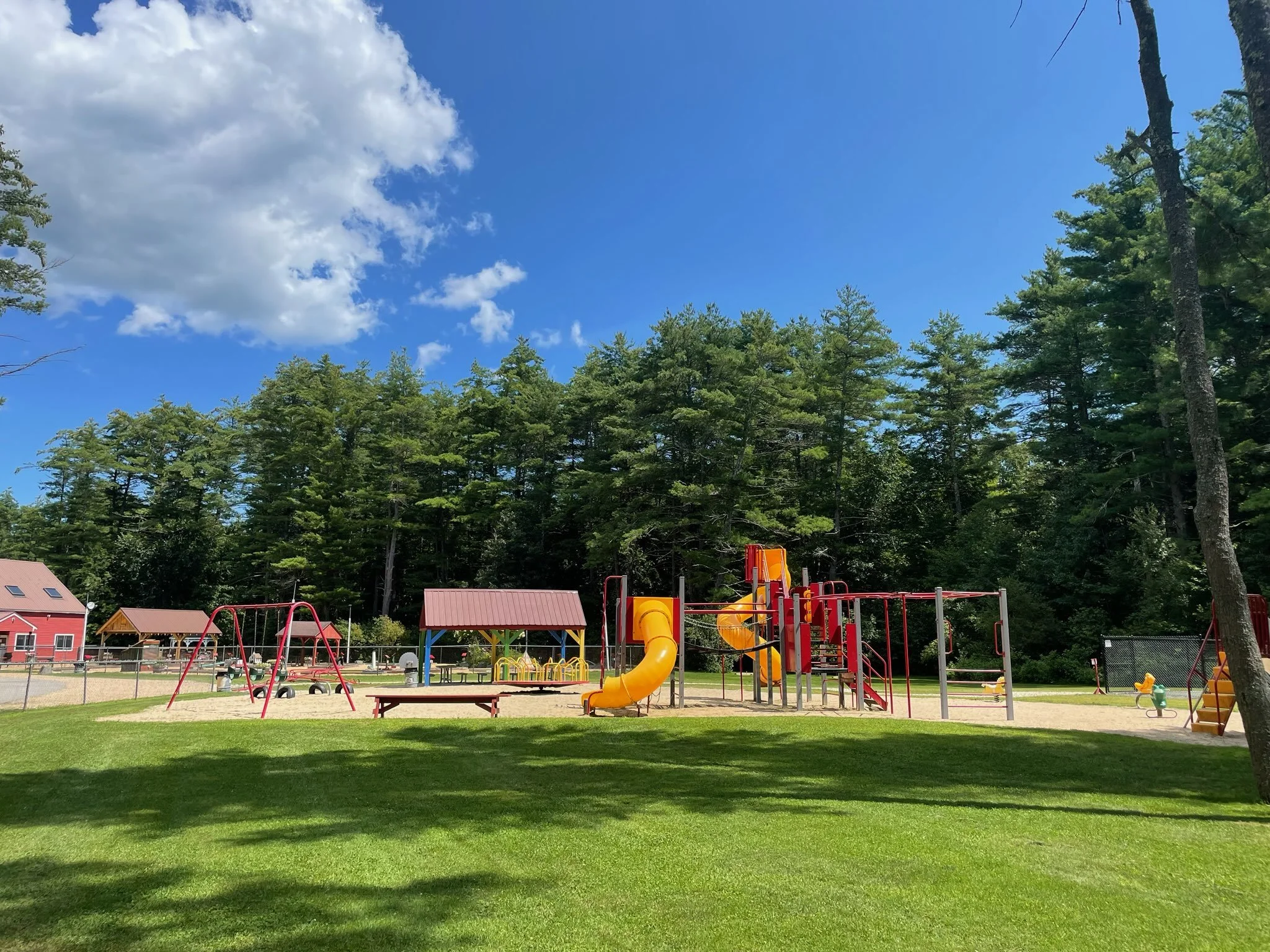 Playground with swings, slides, and a sandbox, surrounded by grass, trees, and a blue sky with clouds.