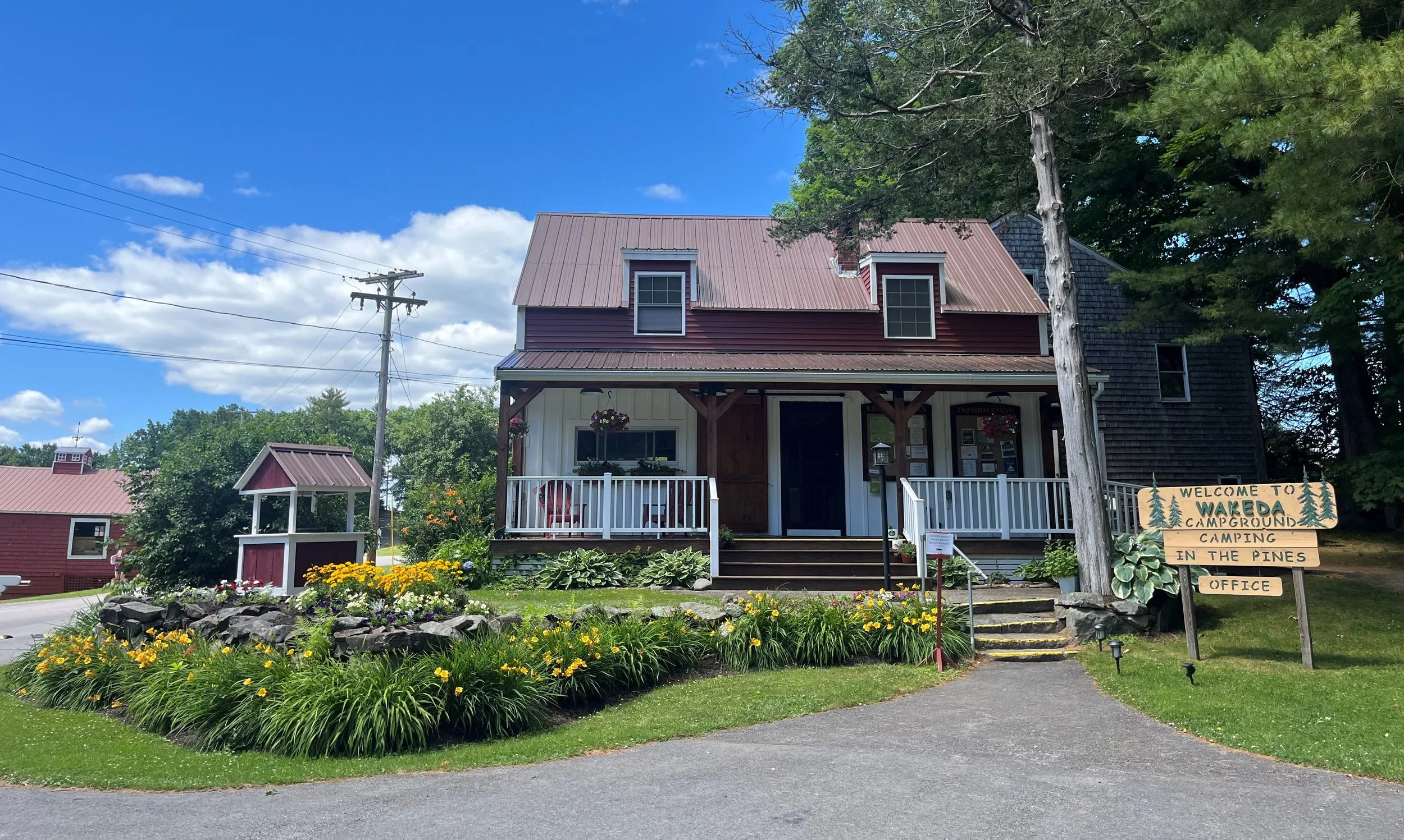 A small countryside building with a porch, surrounded by flower beds and greenery, with a sign that reads 'Welcome to Wareda Campground, Camping in the Pines, Office' under a partly cloudy sky.