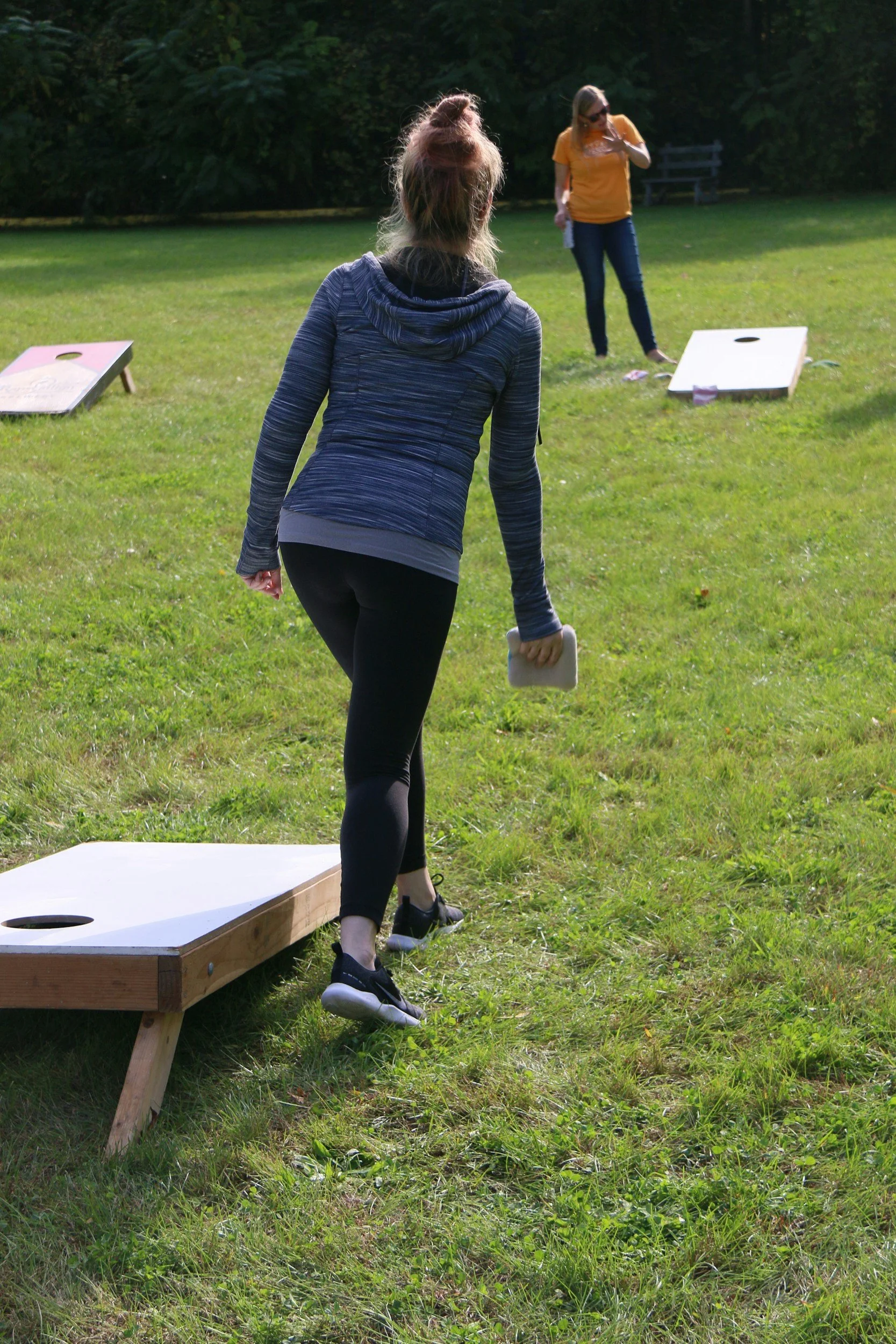 Two women playing cornhole outdoors on a grassy field with trees in the background. One woman is preparing to toss a bean bag, while the other stands near her.
