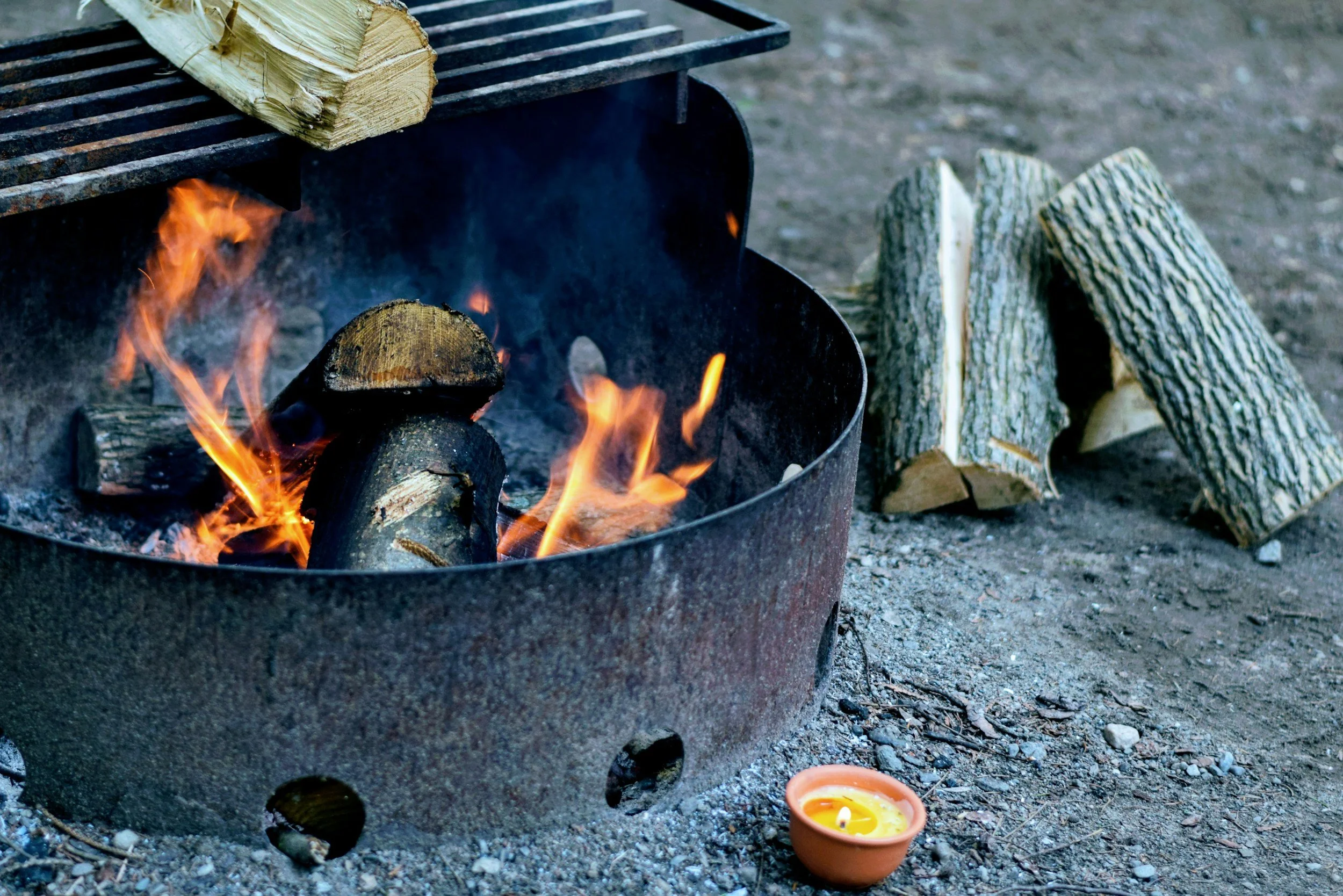 A campfire with burning logs inside a metal fire ring, with logs placed nearby, on dirt ground.