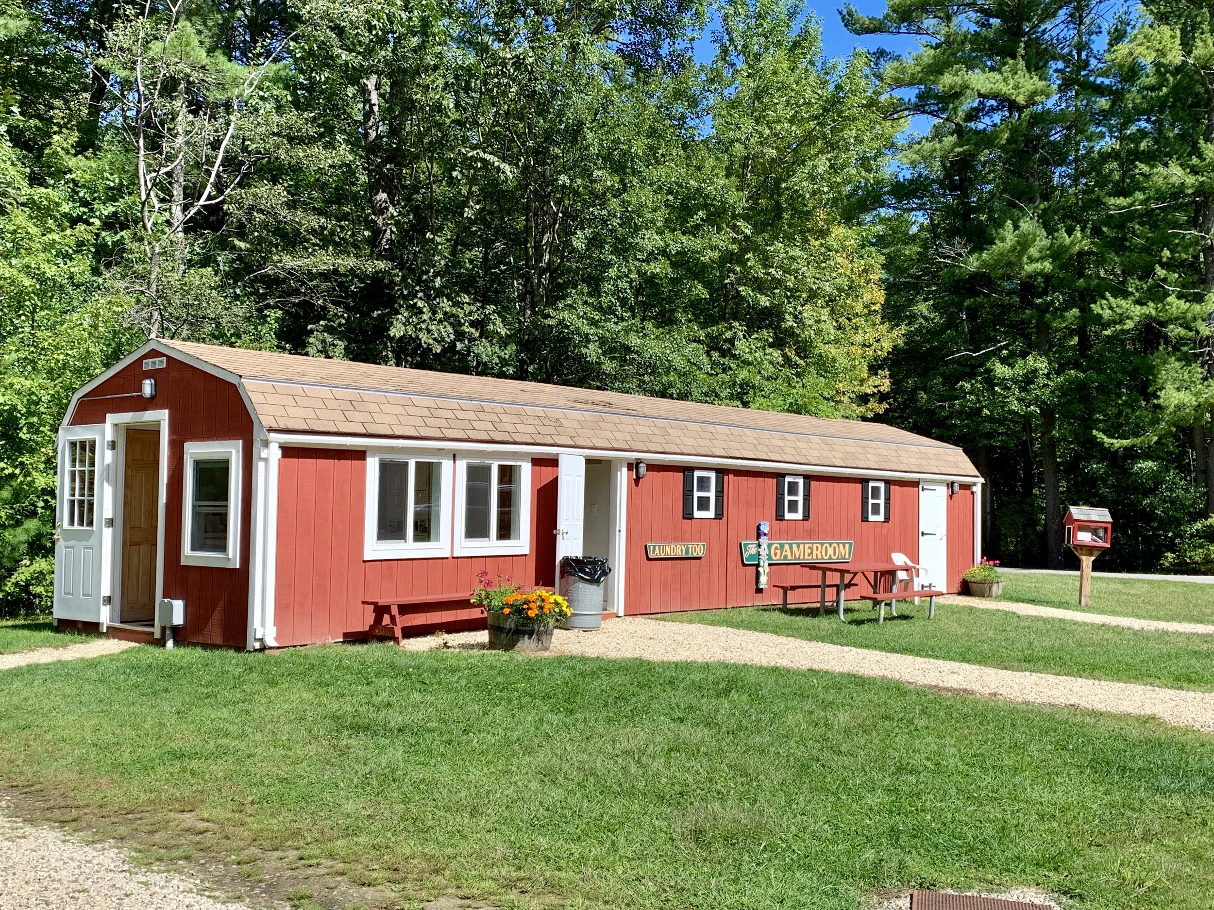 A red building with white trim and a tan roof, surrounded by green grass and trees, with signs reading 'Laundry Too' and 'GAMEROOM', a windowed entrance, outdoor picnic table, and a red mailbox.