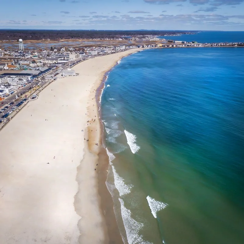 Aerial view of a sandy beach alongside a city, with buildings and parking lot near the coast, and ocean waves approaching the shoreline under a partly cloudy sky.