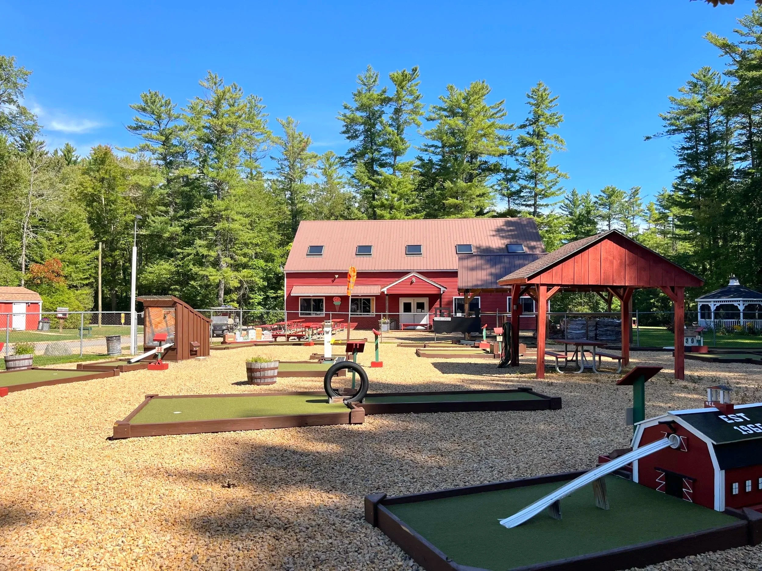 Miniature golf course with multiple holes, surrounded by greenery and trees, under a bright blue sky with some clouds.