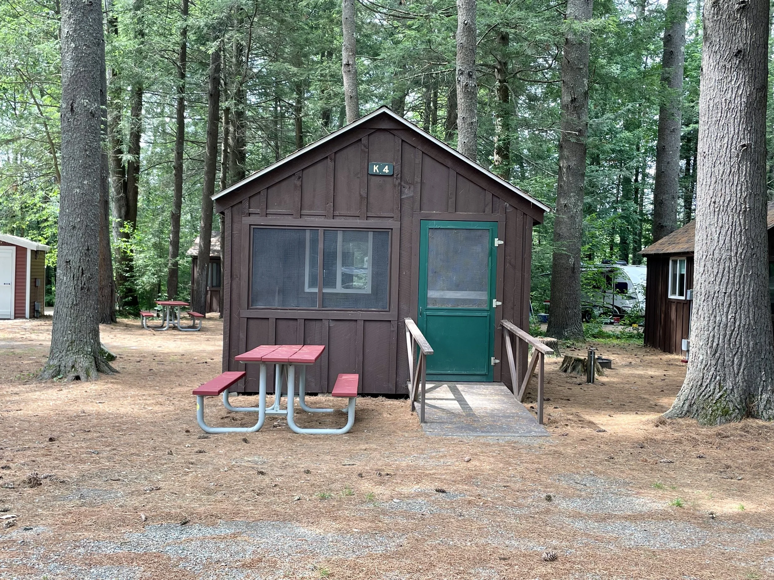 A small brown cabin with a green door and window bars, set in a wooded campsite with tall trees. There are two red picnic tables in front of the cabin, and a small ramp leading to the door. The ground is covered with pine needles and dirt.