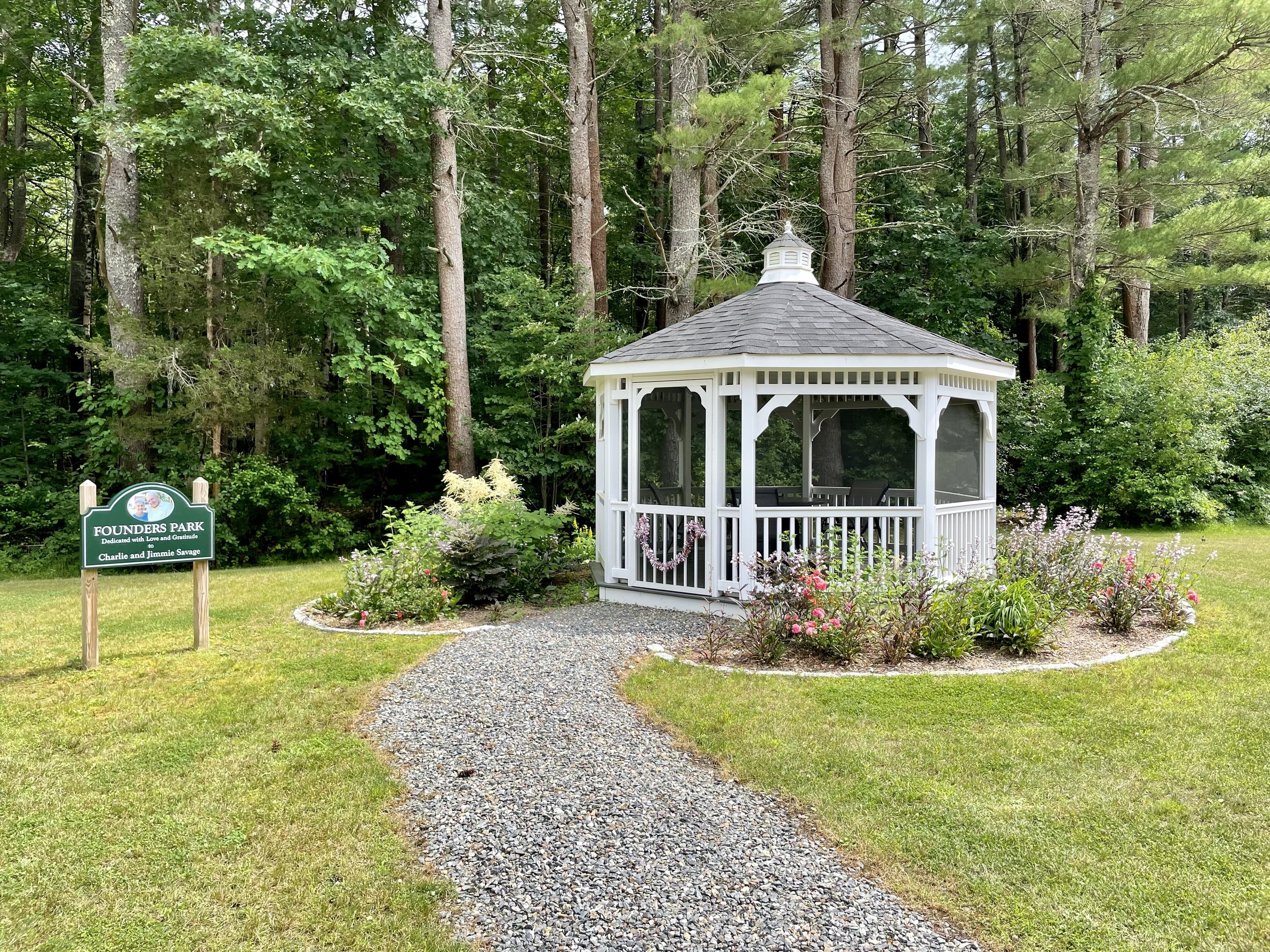 A white gazebo with a shingled roof, surrounded by a flower bed with pink and purple flowers, located in Founders Park with a gravel pathway leading to it and a green park sign nearby in front of a wooded background.
