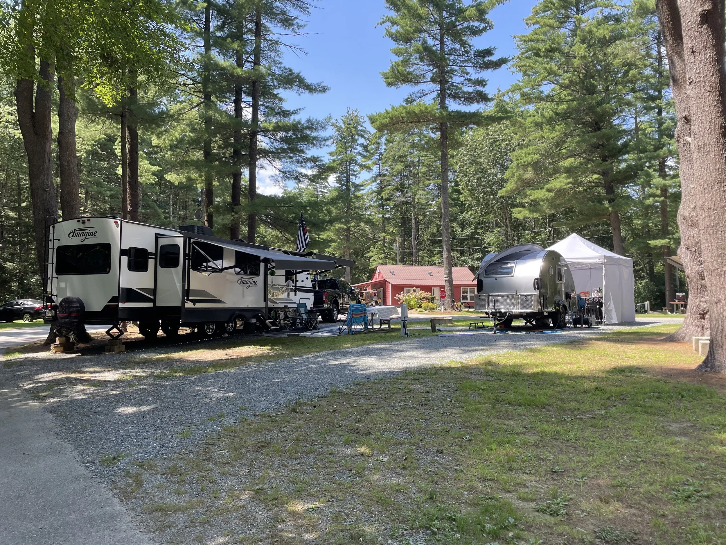 A campground with two RV trailers set up among tall trees. The first trailer is larger with a black and white exterior, an awning, and an American flag. The second is a smaller silver Airstream-style trailer with an attached white canopy. Surrounding