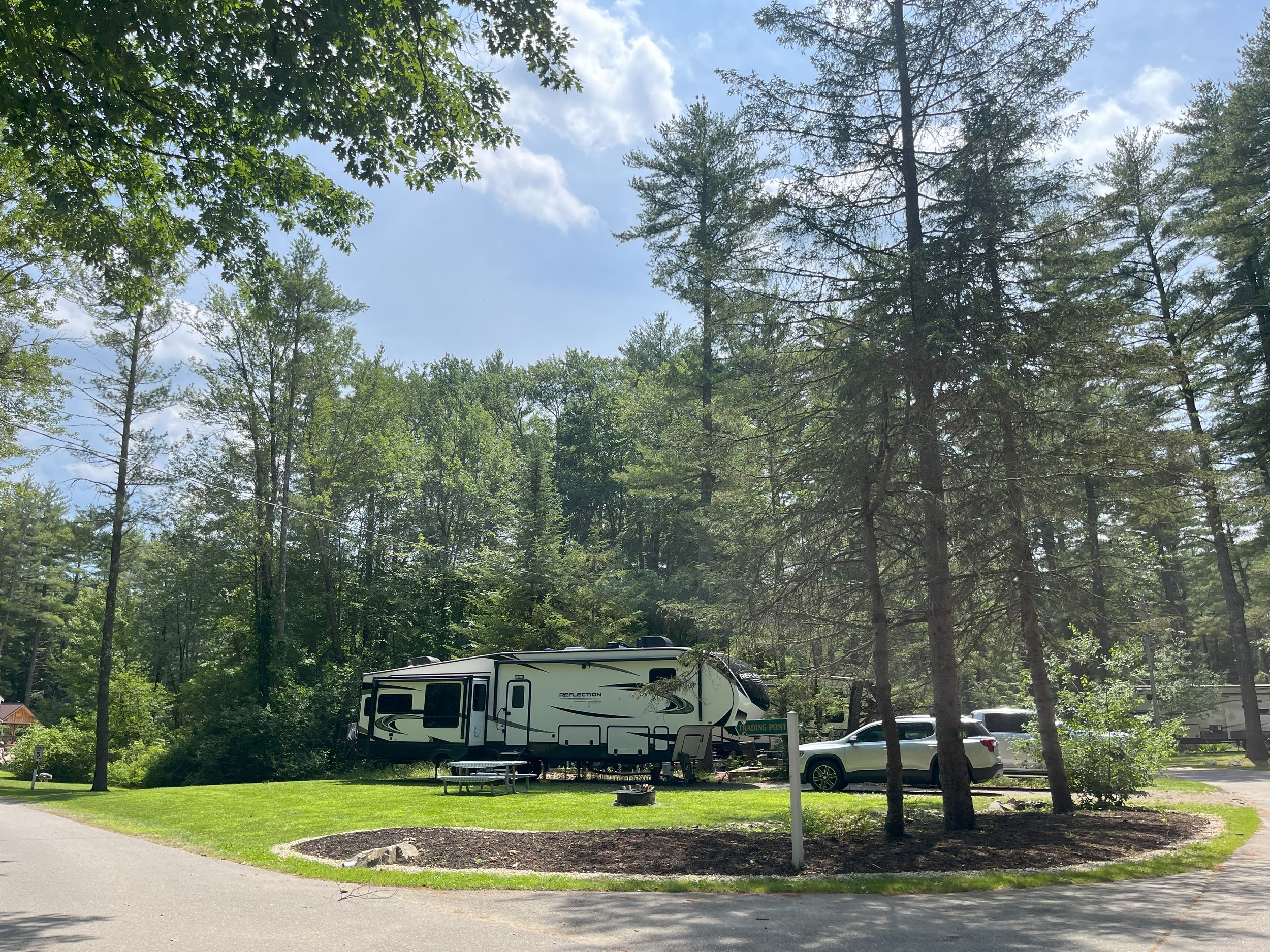 A recreational vehicle trailer parked in a grassy camping site surrounded by tall trees, with a silver car parked beside it. The sky is blue with some clouds, and there is a reading post sign in front of the trailer.