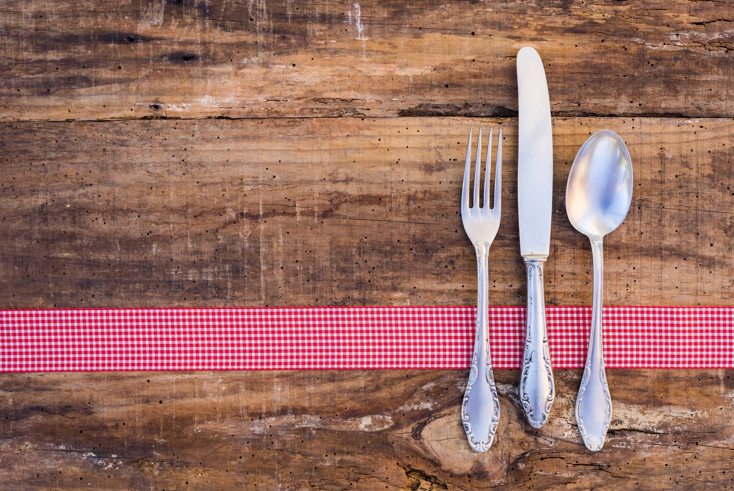 Silver fork, knife, and spoon placed on a rustic wooden table with a red and white checkered strip running horizontally across the bottom of the image.