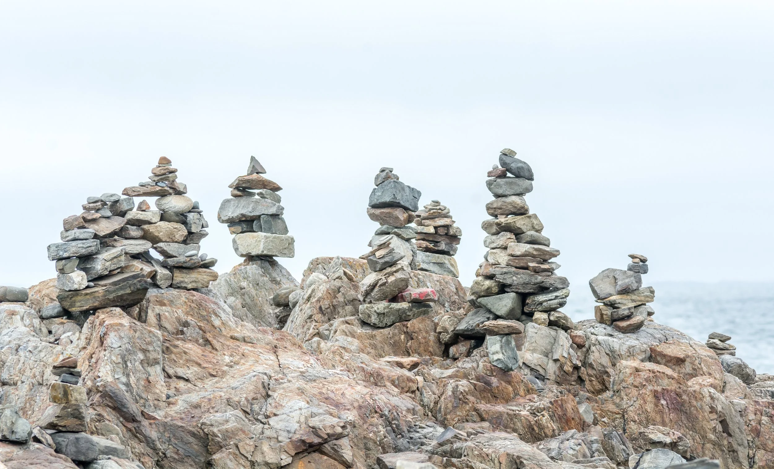 Multiple stacks of balanced rocks arranged on a rocky shoreline, with a cloudy sky and the sea in the background.