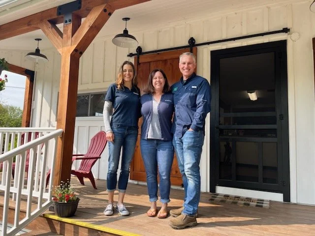Three people standing together on a wooden porch, smiling at the camera. There are two women and one man, dressed casually in jeans and shirts, with a building and porch furniture visible behind them.