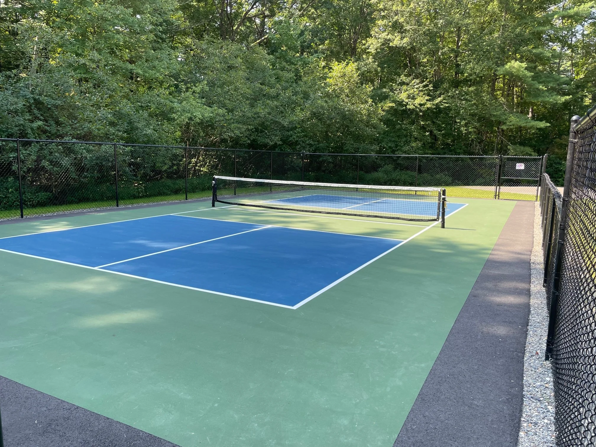 Empty outdoor tennis court with green and blue surface, surrounded by a black fence, set against a backdrop of trees and greenery.