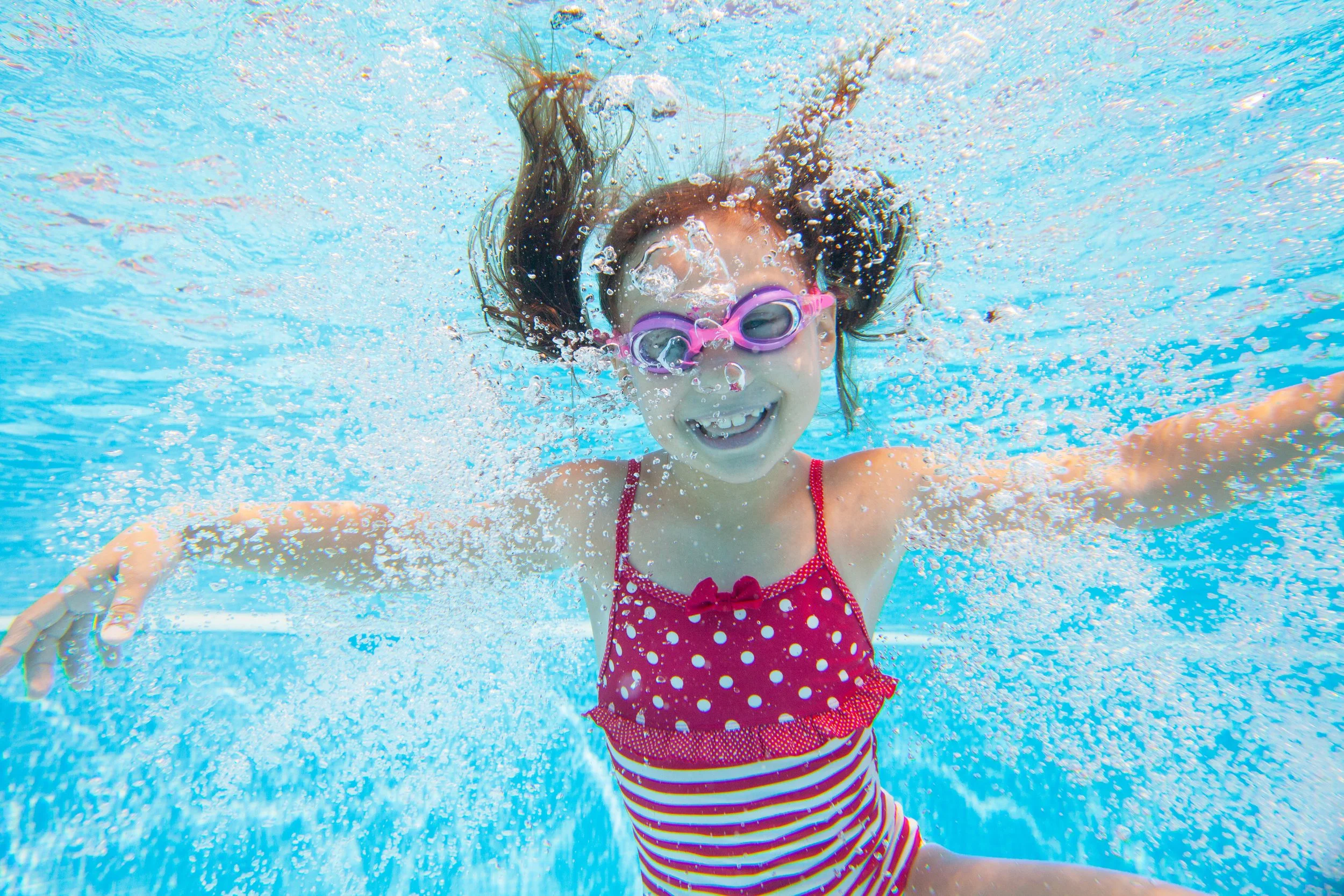 A young girl swimming underwater in a pool, wearing pink goggles and a red polka-dot swimsuit, smiling with her arms spread out.