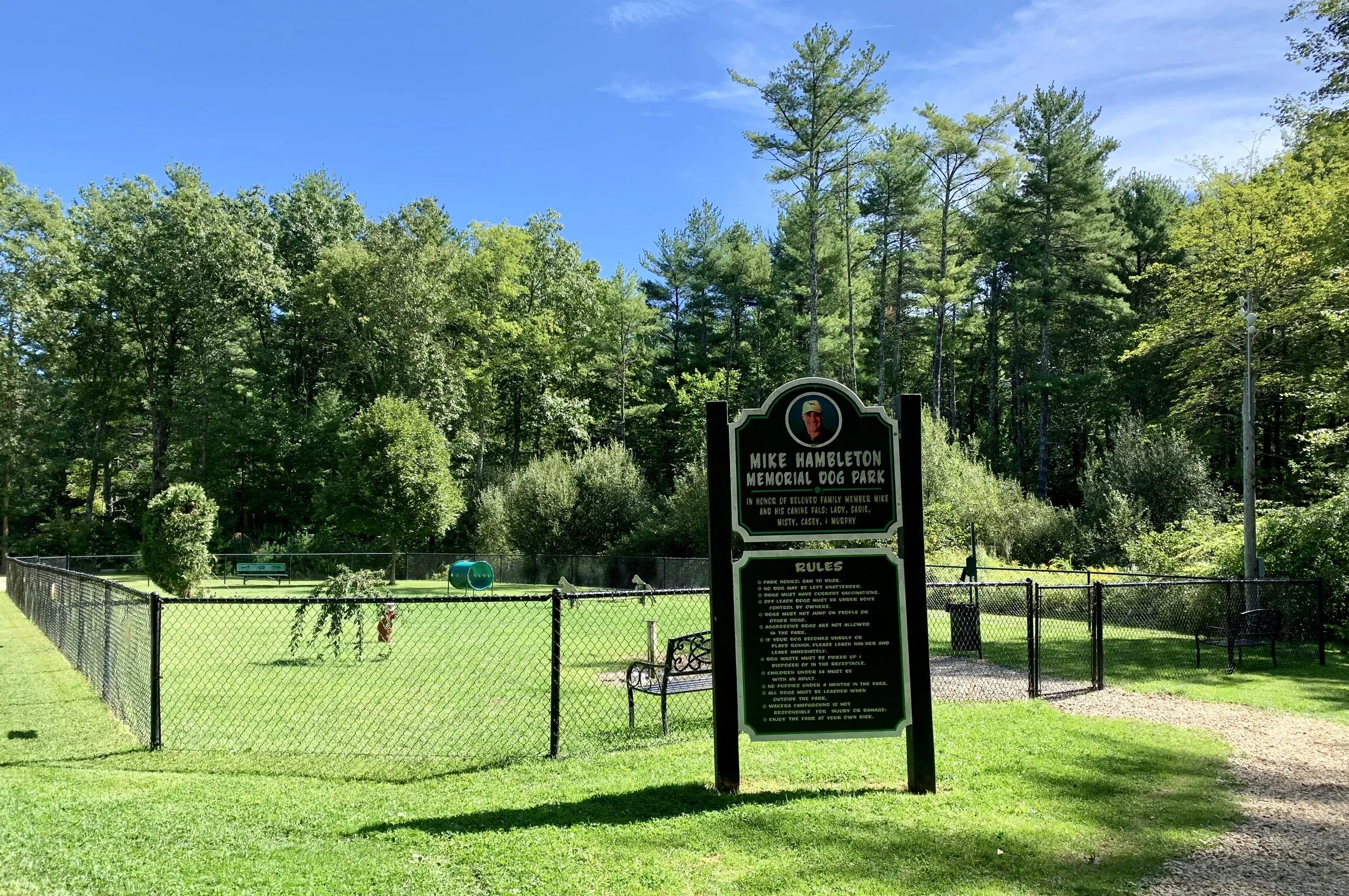 Signboard at Mike Hambleton Memorial Dog Park with rules and photo of Mike Hambleton, surrounded by green grass, trees, and blue sky.