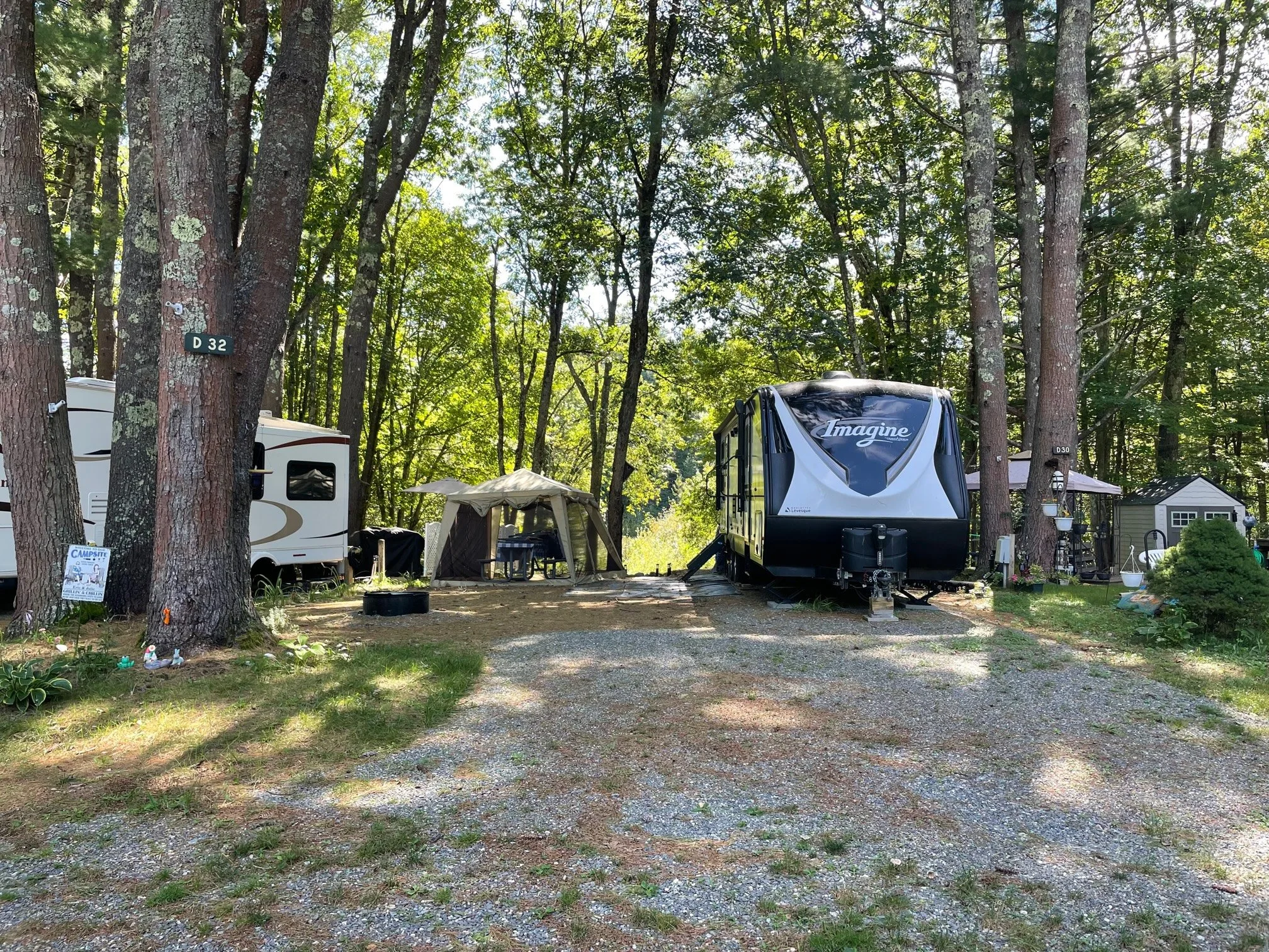 A camping site in a wooded area with two RVs, a tent, a table with an umbrella, and various camping gear.