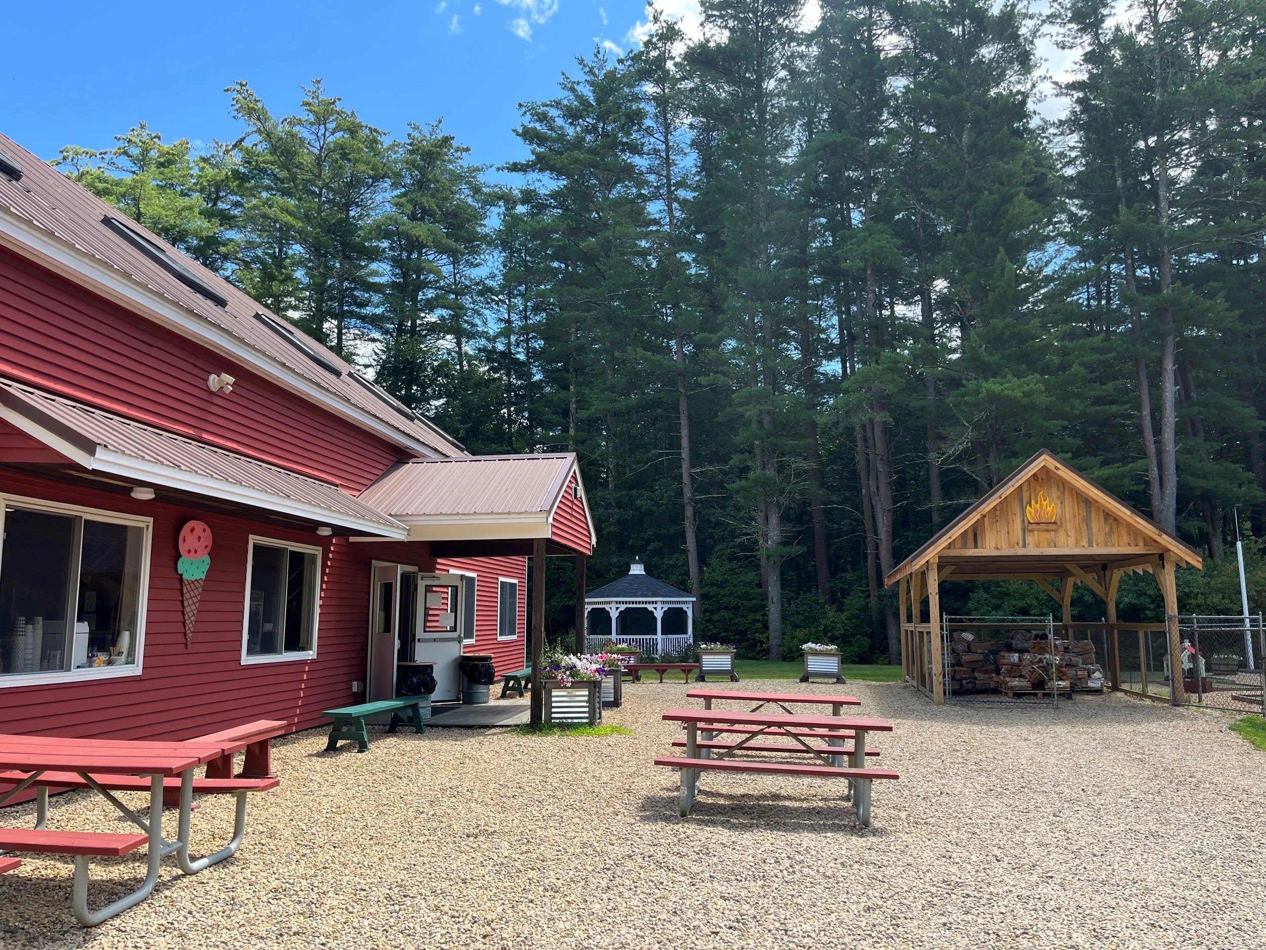 Red building with ice cream cone decoration, outdoor seating with picnic tables, a wooden shelter with logs, a gazebo, and a forested background.