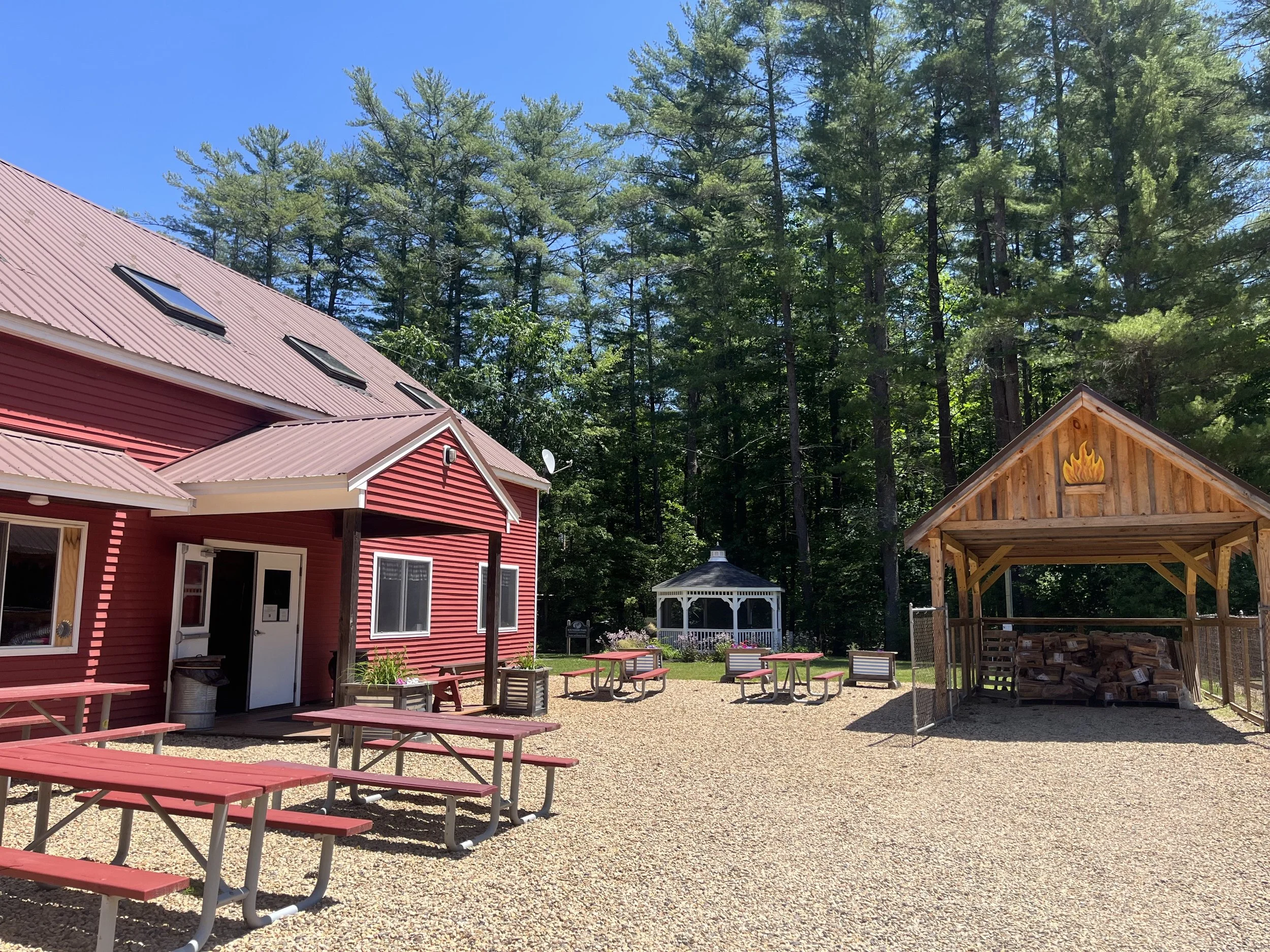 A red building with ice cream cone decoration, tall pine trees in the background, there are picnic tables, a gazebo, and a woodpile shelter in an outdoor area with gravel ground.