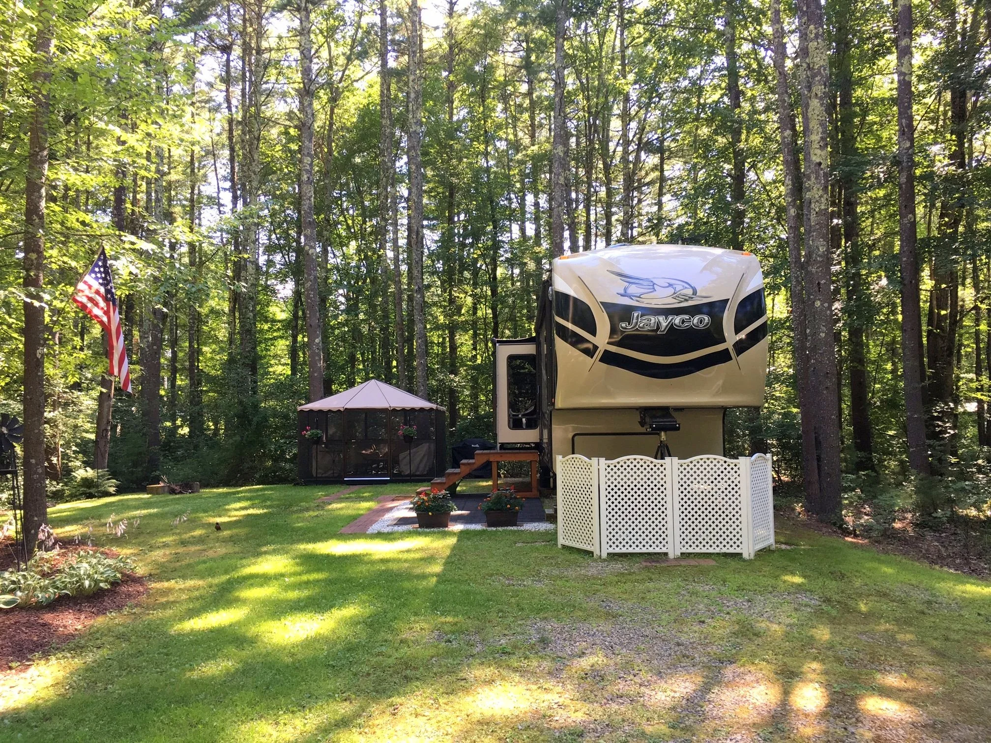 A backyard with a large RV trailer, a white picket fence, a small shed, potted plants, an American flag, and trees in the background.