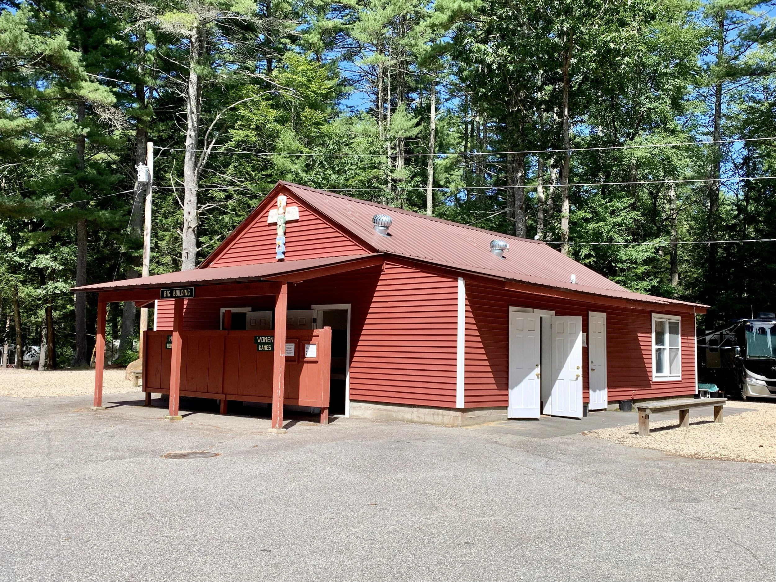 Red building with a brown metal roof, surrounded by trees with a parking lot in front. Sign indicates it is a bathroom, with separate men's and women's entrances.
