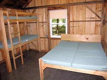 Interior of a wooden room with a bunk bed and a double bed, both with blue mattresses, and a window with a view of trees outside.