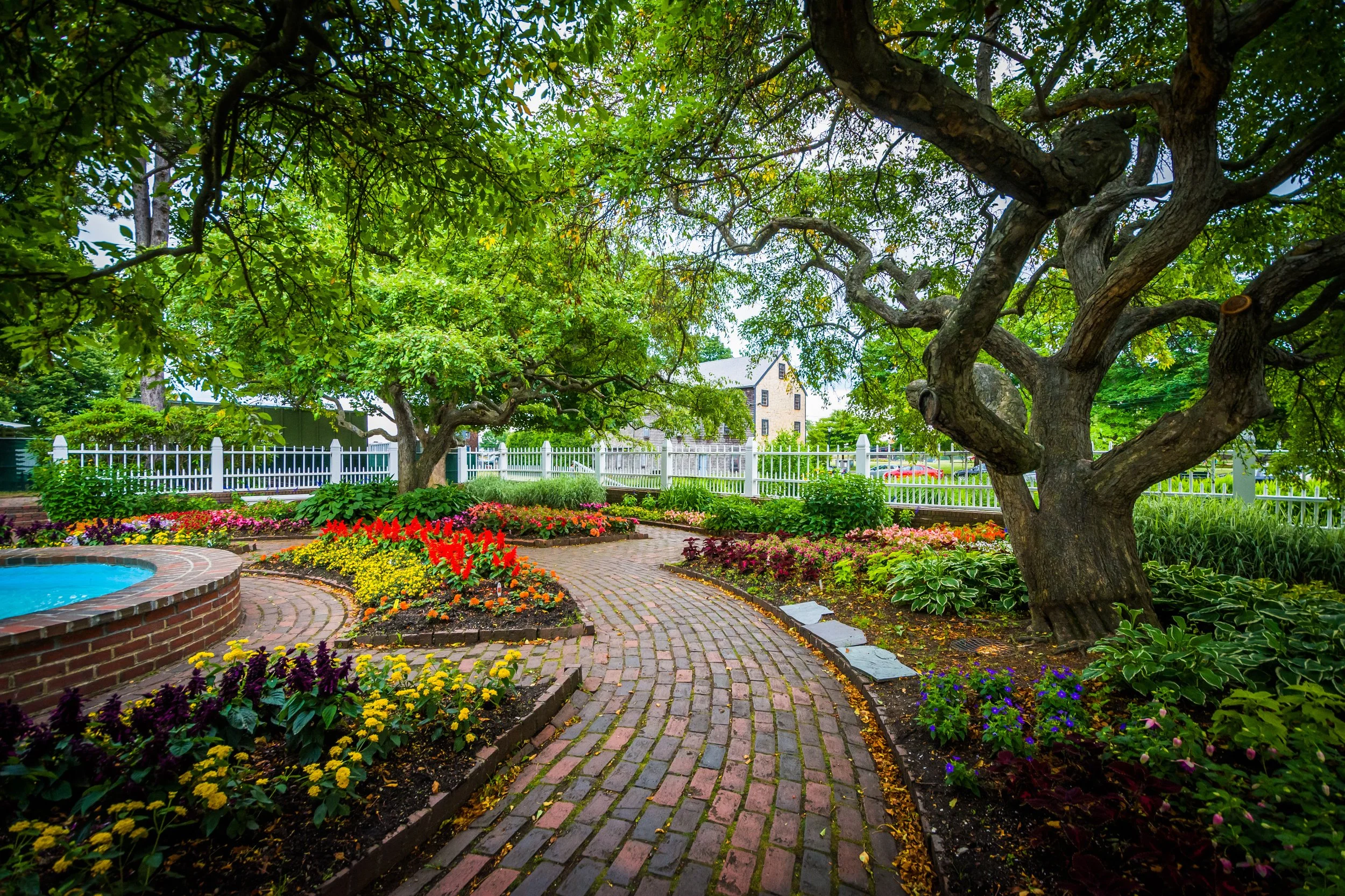 A garden with a brick pathway, colorful flowers, large green trees, and a white fence.
