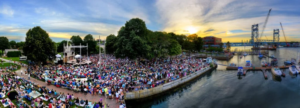 Large outdoor concert at sunset by the river with a big crowd, trees, and boats docked along the waterfront.