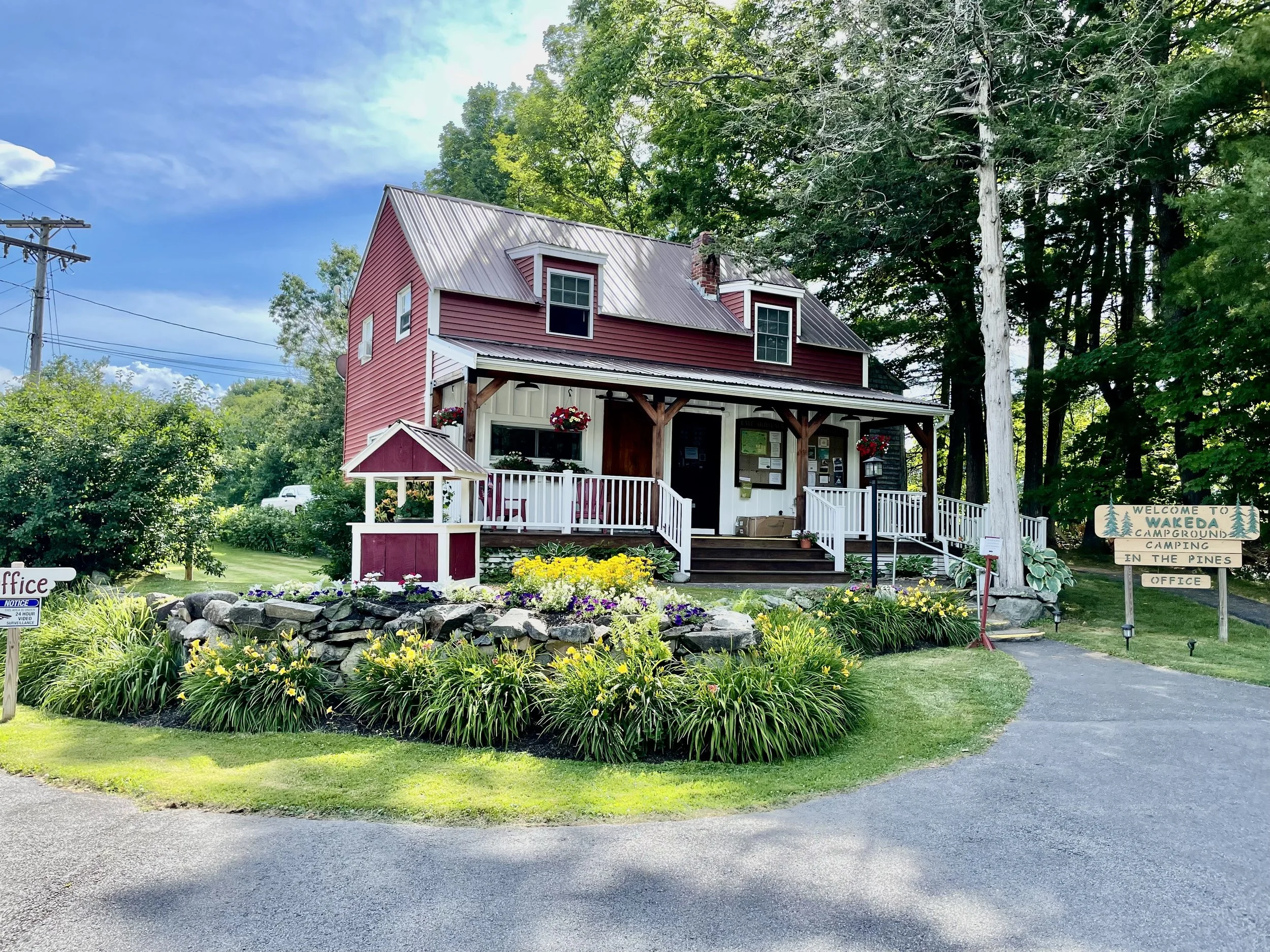 A red and white office with a porch, surrounded by green plants and flowers, and a sign to a campground in a wooded area.
