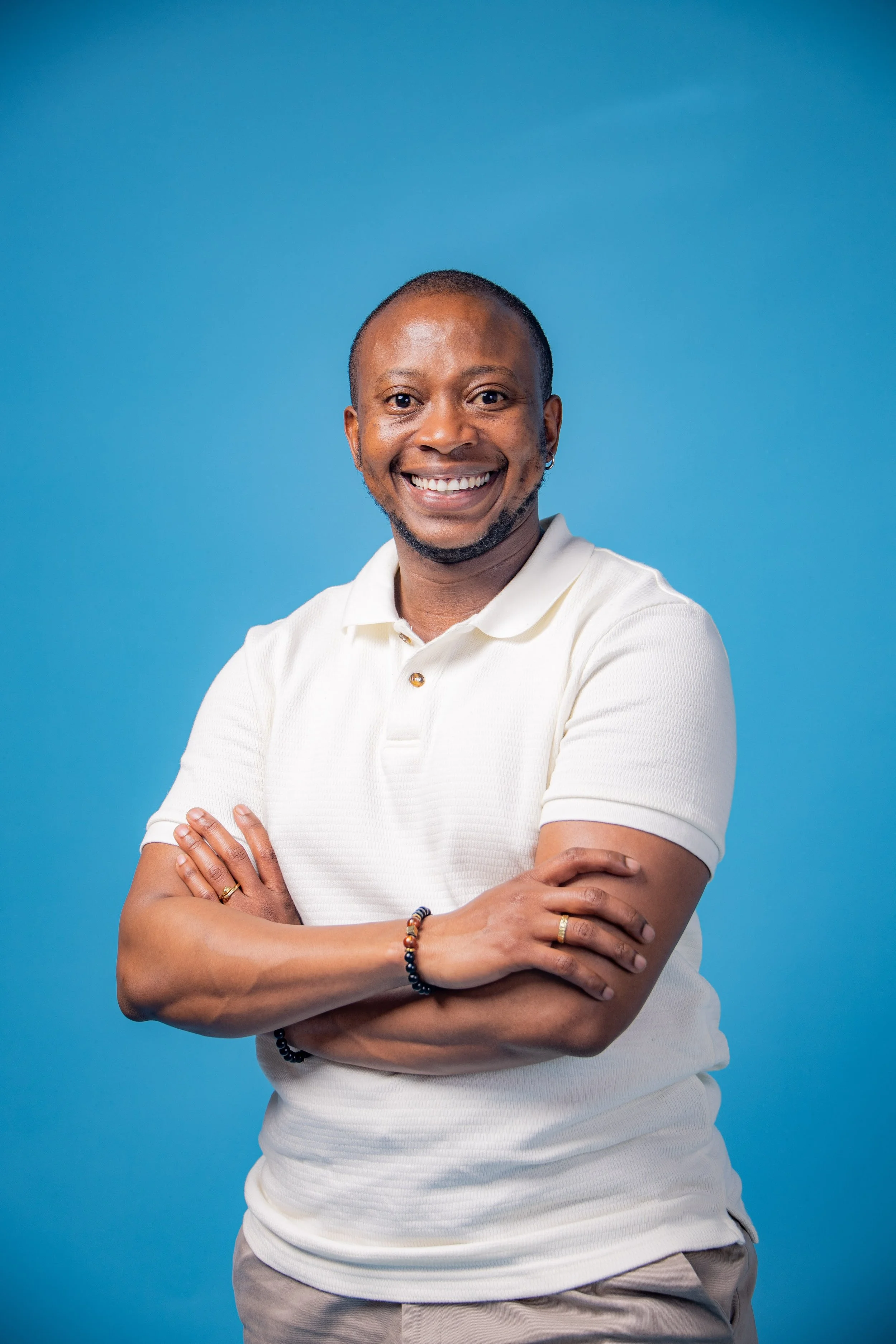 A smiling man with short hair and a beard, wearing a white polo shirt, standing with arms crossed against a blue background.