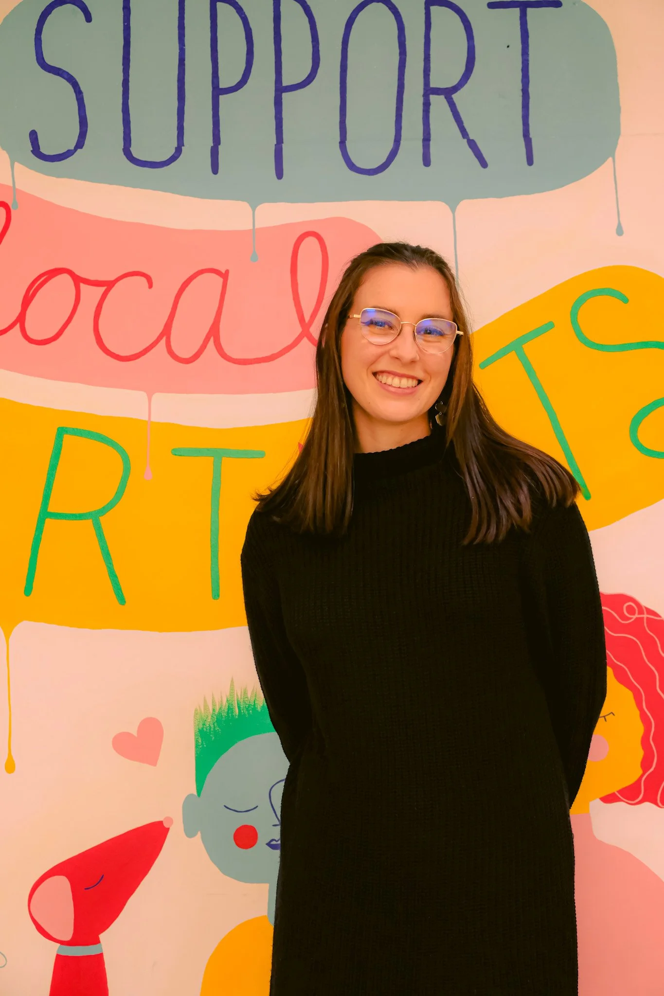 A young woman with glasses and long brown hair smiling in front of a colorful mural that reads support local artists.