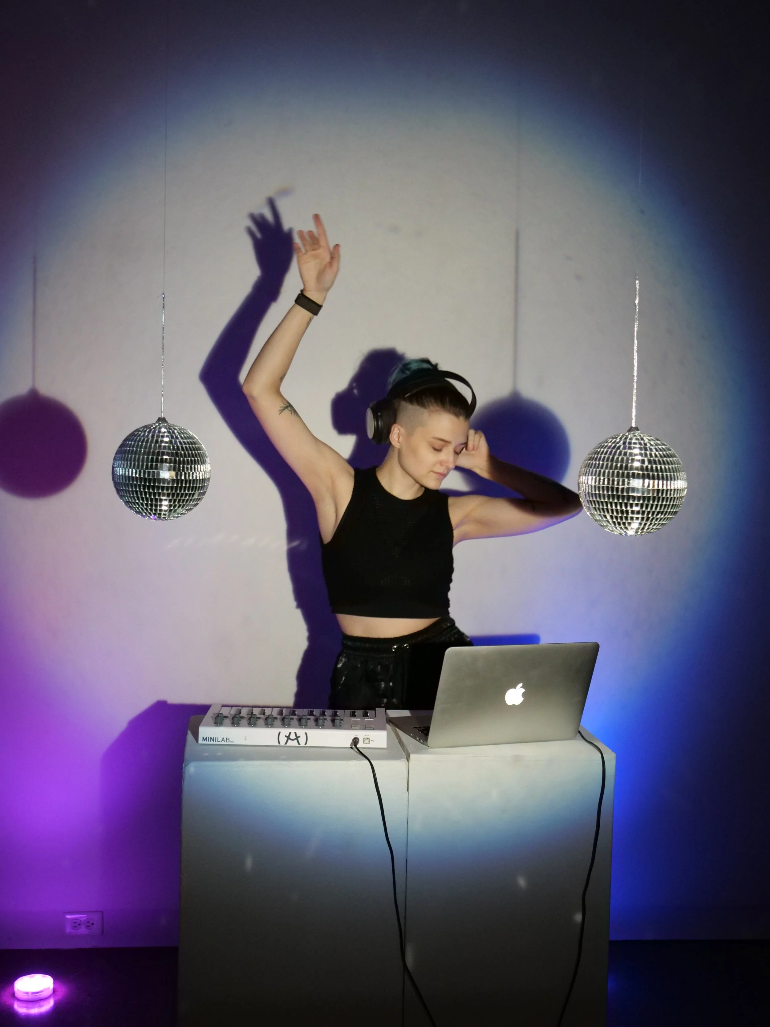 A woman with short hair, wearing headphones and a black crop top, is dancing with one arm raised and one arm bent, surrounded by disco balls, with DJ equipment and a laptop on a white stand in front of her. Shadows of the disco balls are cast on the wall behind her.