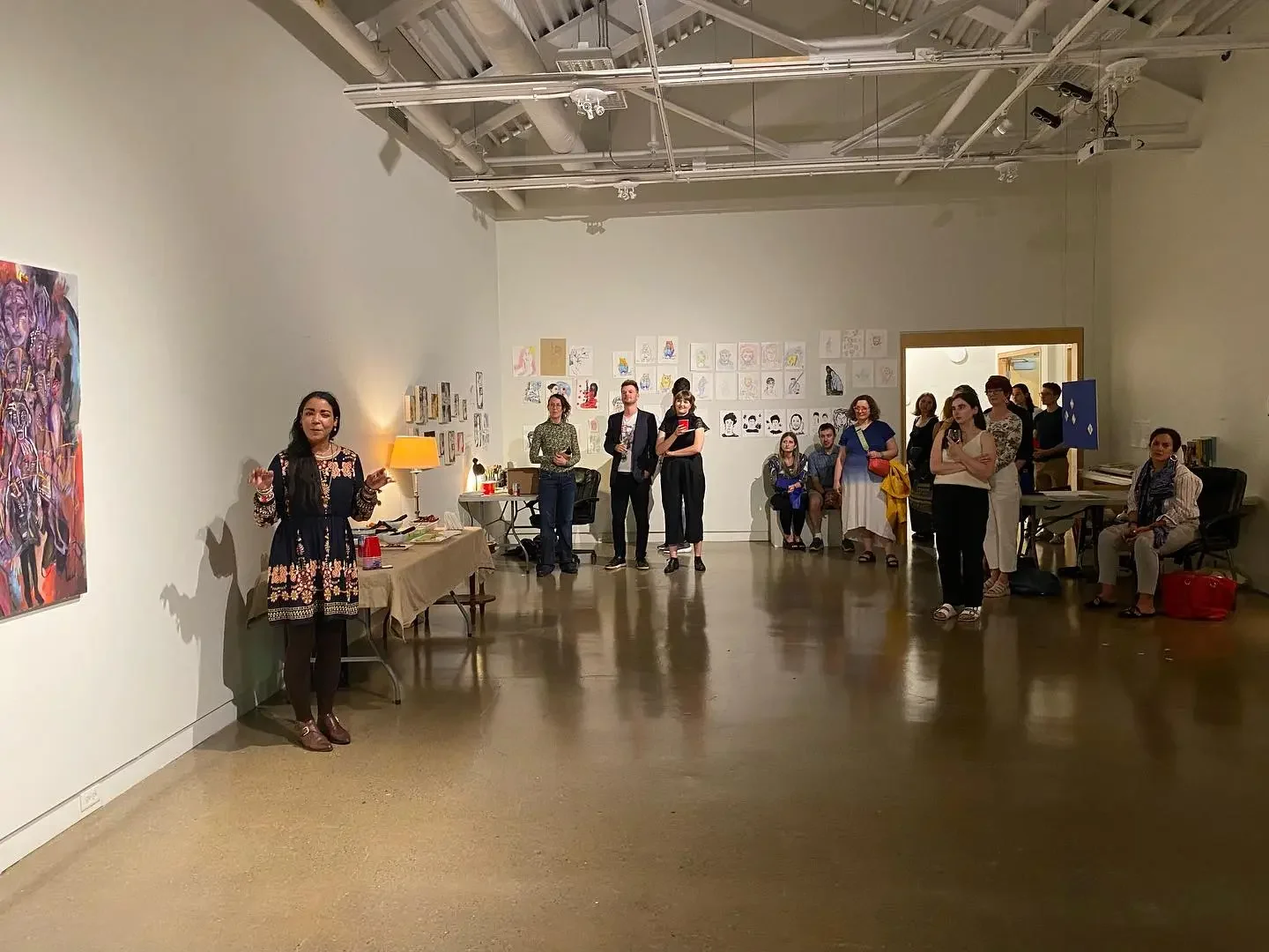 A group of people attending an art gallery opening, with some listening to a woman speaking, and artwork displayed on the walls.