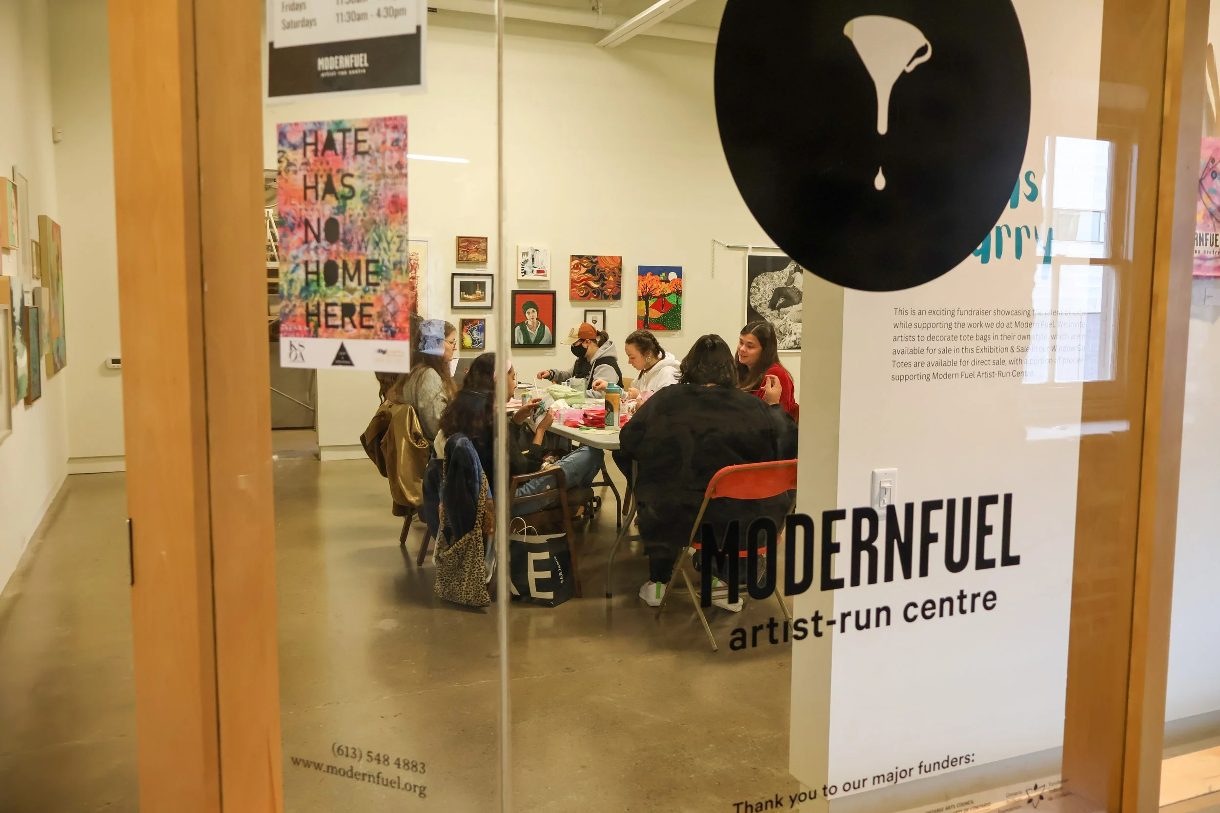 People sitting at a table in Modern Fuel Artist-Run Centre, with artwork on the walls behind them, seen through a glass door with a poster and sticker on it.