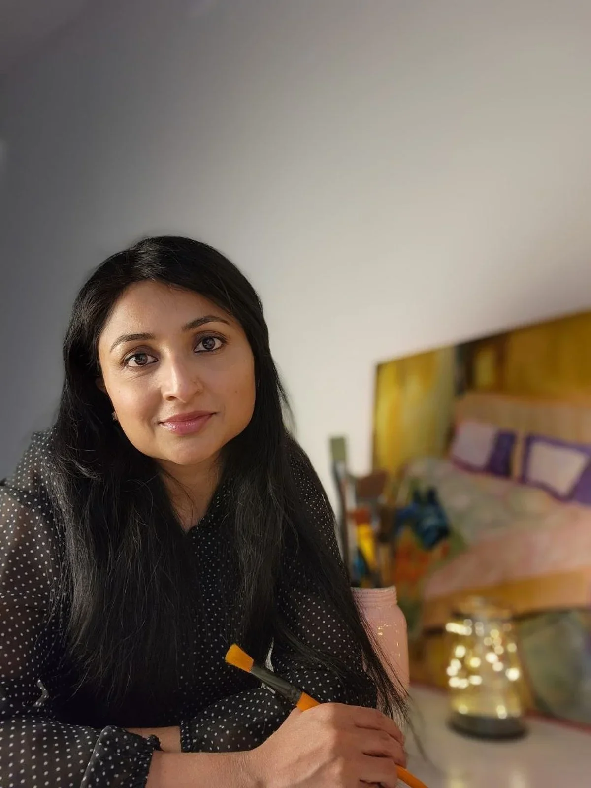 A woman with long black hair and a black polka dot blouse sitting at a table with a pink cup and pen. There is a bed with purple pillows and a yellow headboard in the background.
