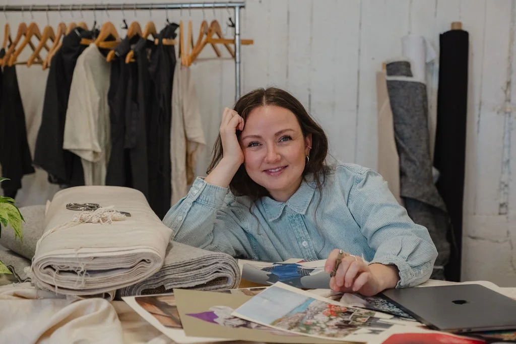 A woman sitting at a table surrounded by fabric swatches and photographs, with clothing on hangers behind her in a workspace.