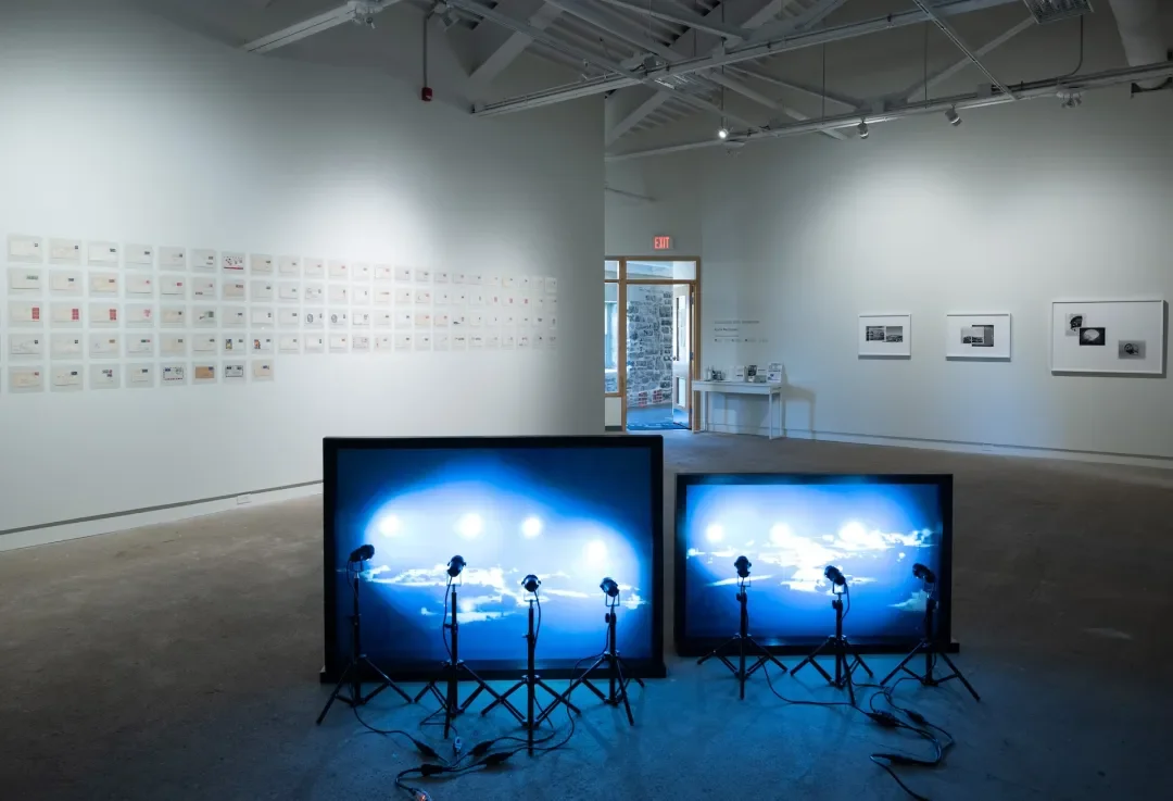 Art gallery with wall displays of small photographs, and two illuminated digital screens with lighting equipment in the foreground.