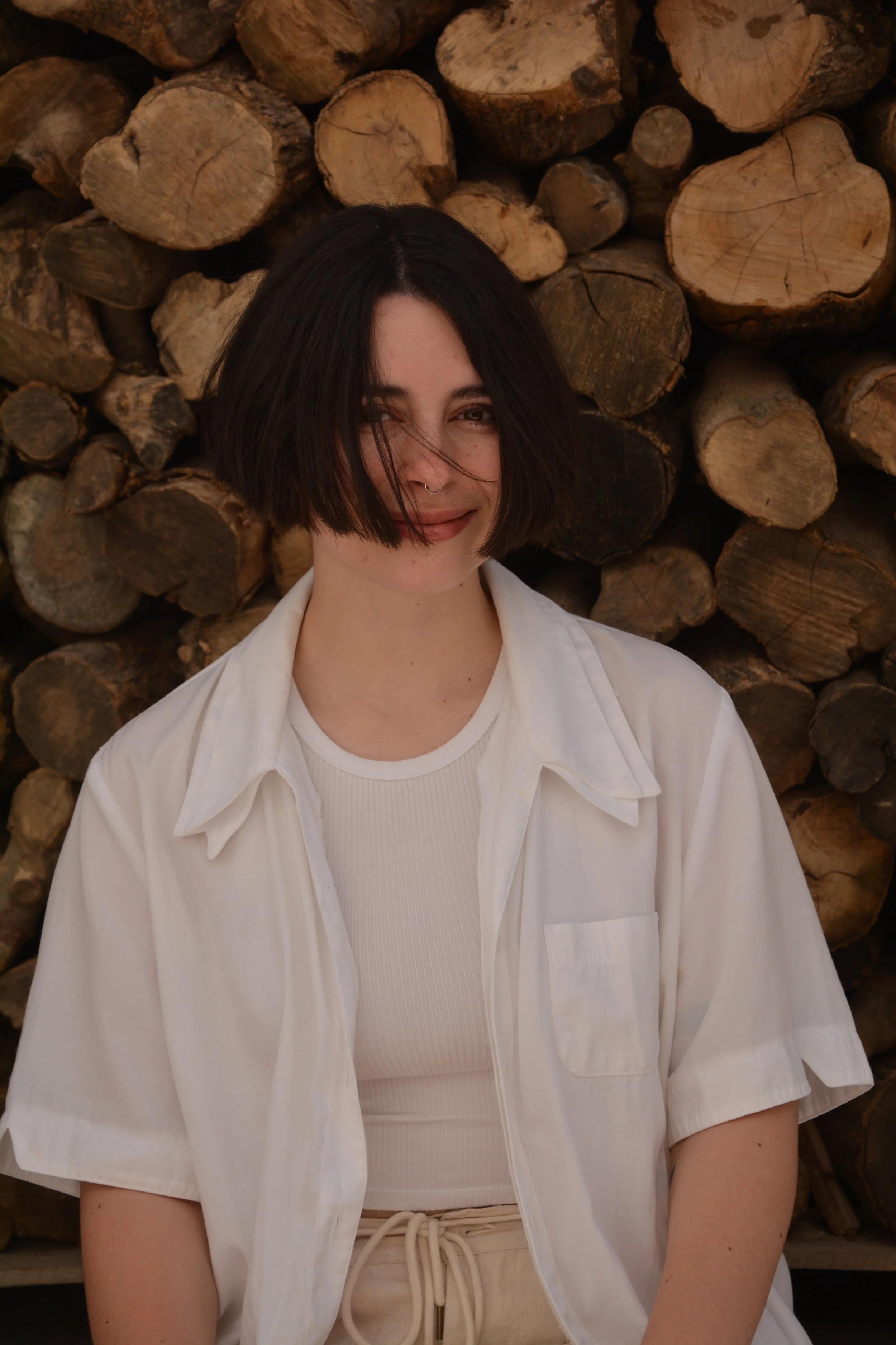 A young woman with short black hair, wearing a white shirt over a white ribbed top, smiling with her eyes slightly squinted, standing in front of a background of stacked firewood.