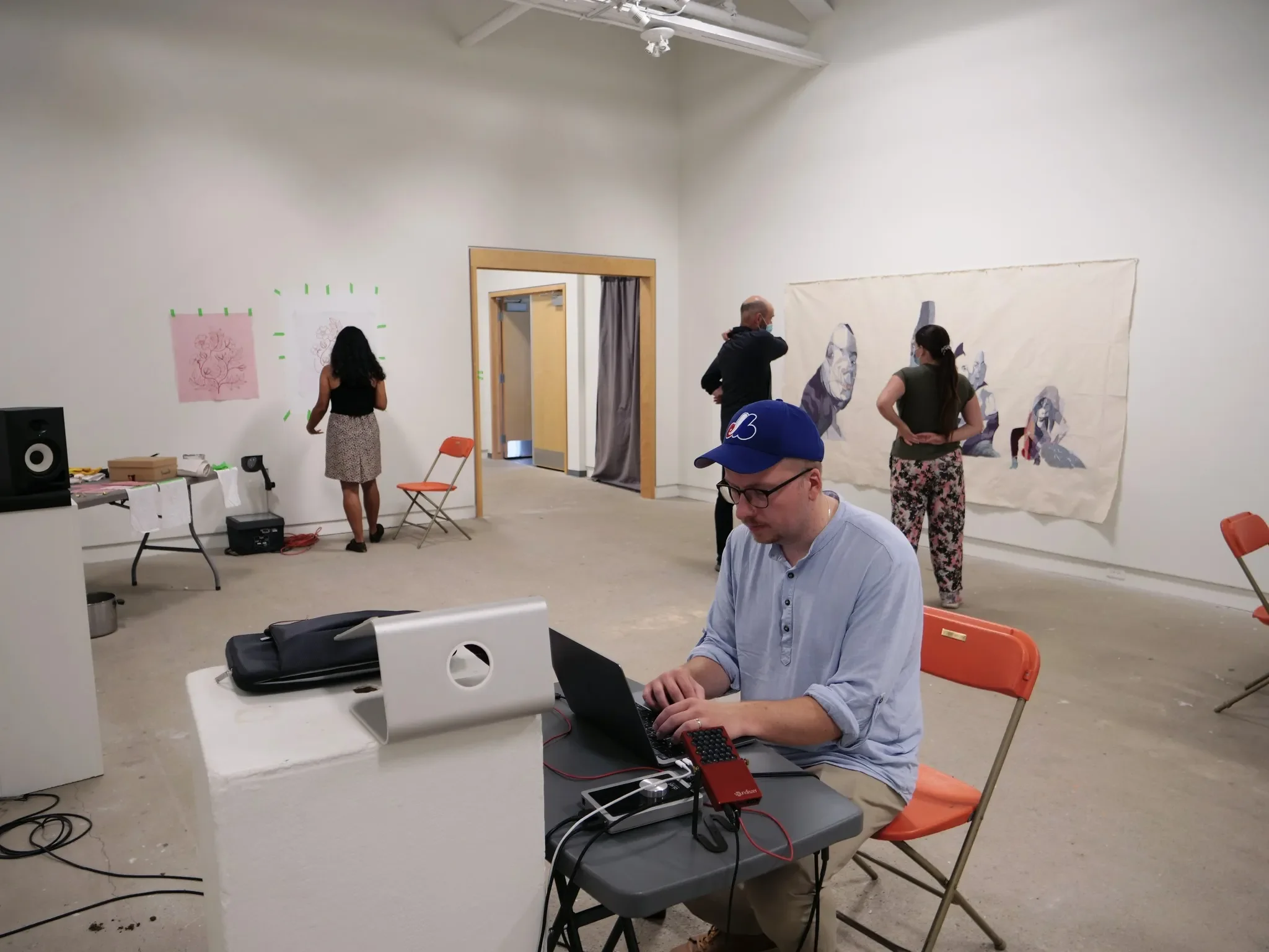 A man wearing glasses and a blue cap working on a laptop at a table, with an art gallery in the background featuring people observing artwork.