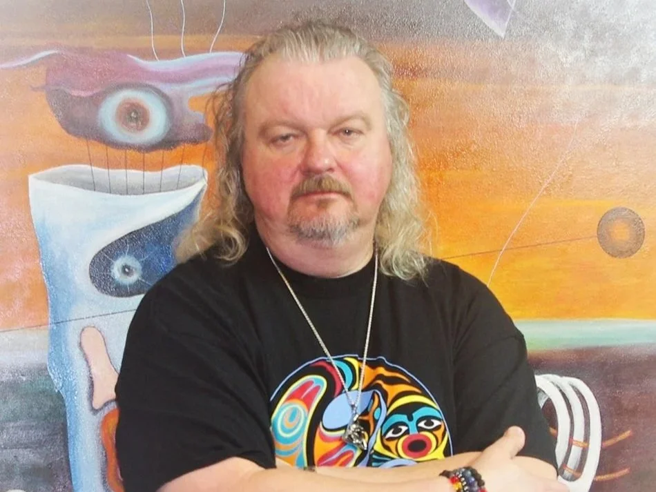 A man with long, curly gray hair and a beard is standing in front of a colorful abstract painting, wearing a black T-shirt with a vibrant, stylized face design and a necklace.