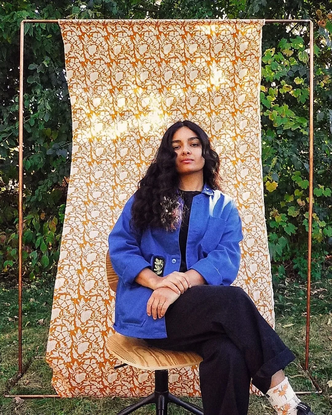 A woman with long curly hair sitting on a wooden chair outside, wearing a blue jacket over a black shirt, with a patterned backdrop and greenery around.