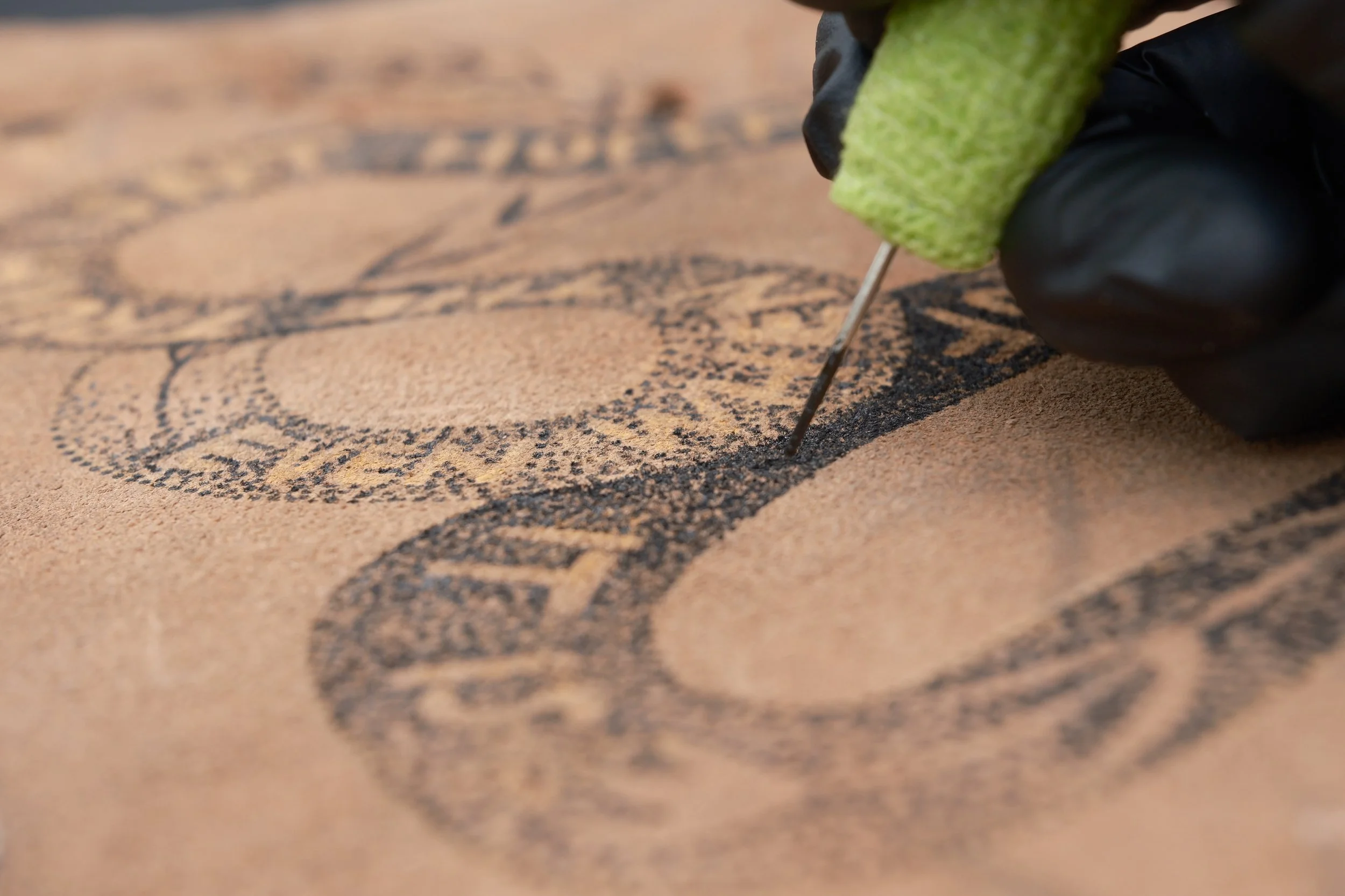 Close-up of a person using a needle and green foam pad to create a stippled design of a face on a piece of leather or paper.
