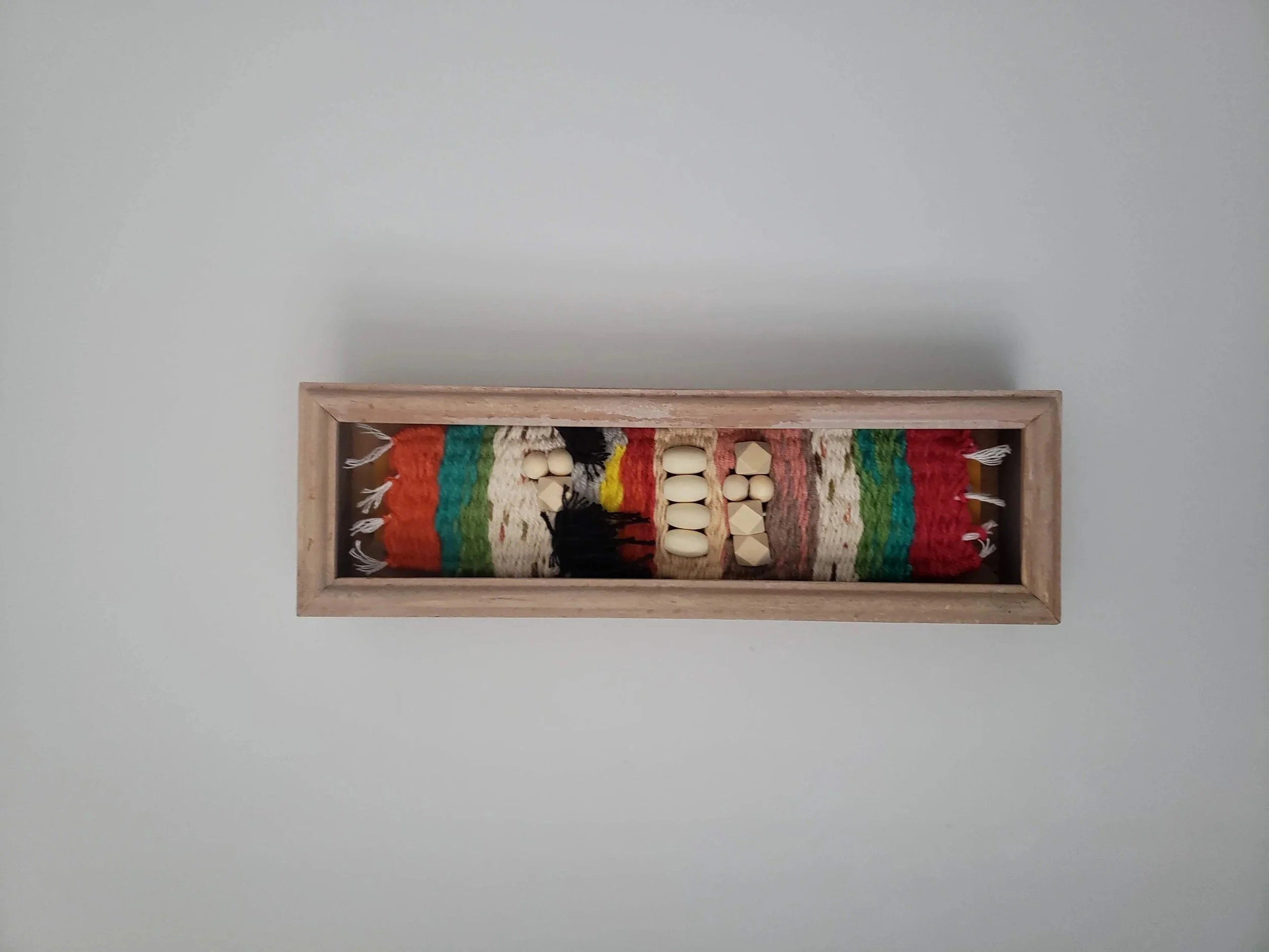 Colorful woven wall hanging with fringe in a wooden frame on a plain gray wall.