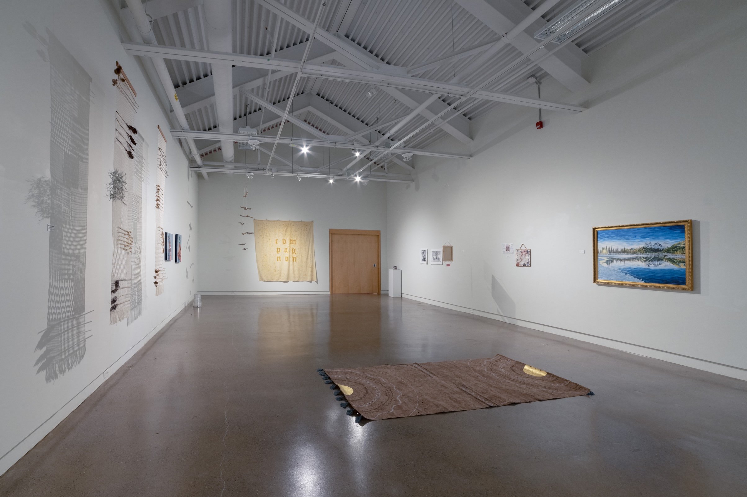 An art gallery with various artworks on white walls, including textiles, paintings, and mixed media pieces. The gallery has a polished concrete floor, high ceiling with exposed beams and spot lighting, and minimal decor with a woven rug on the floor.