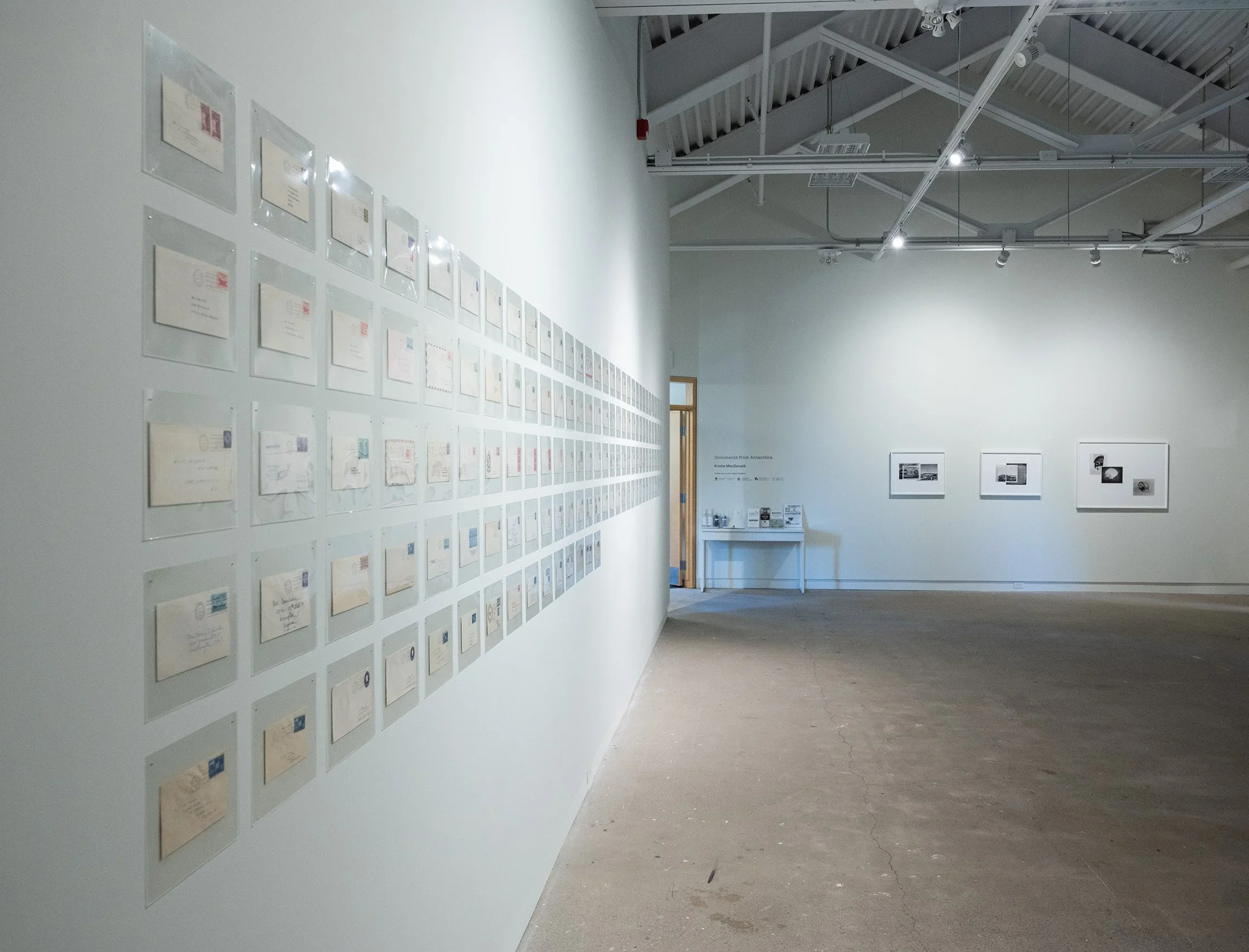 An art gallery with a white wall displaying numerous framed pieces, and three additional framed photographs on the adjacent wall. There is a small table with informational pamphlets near the back, under a sign.