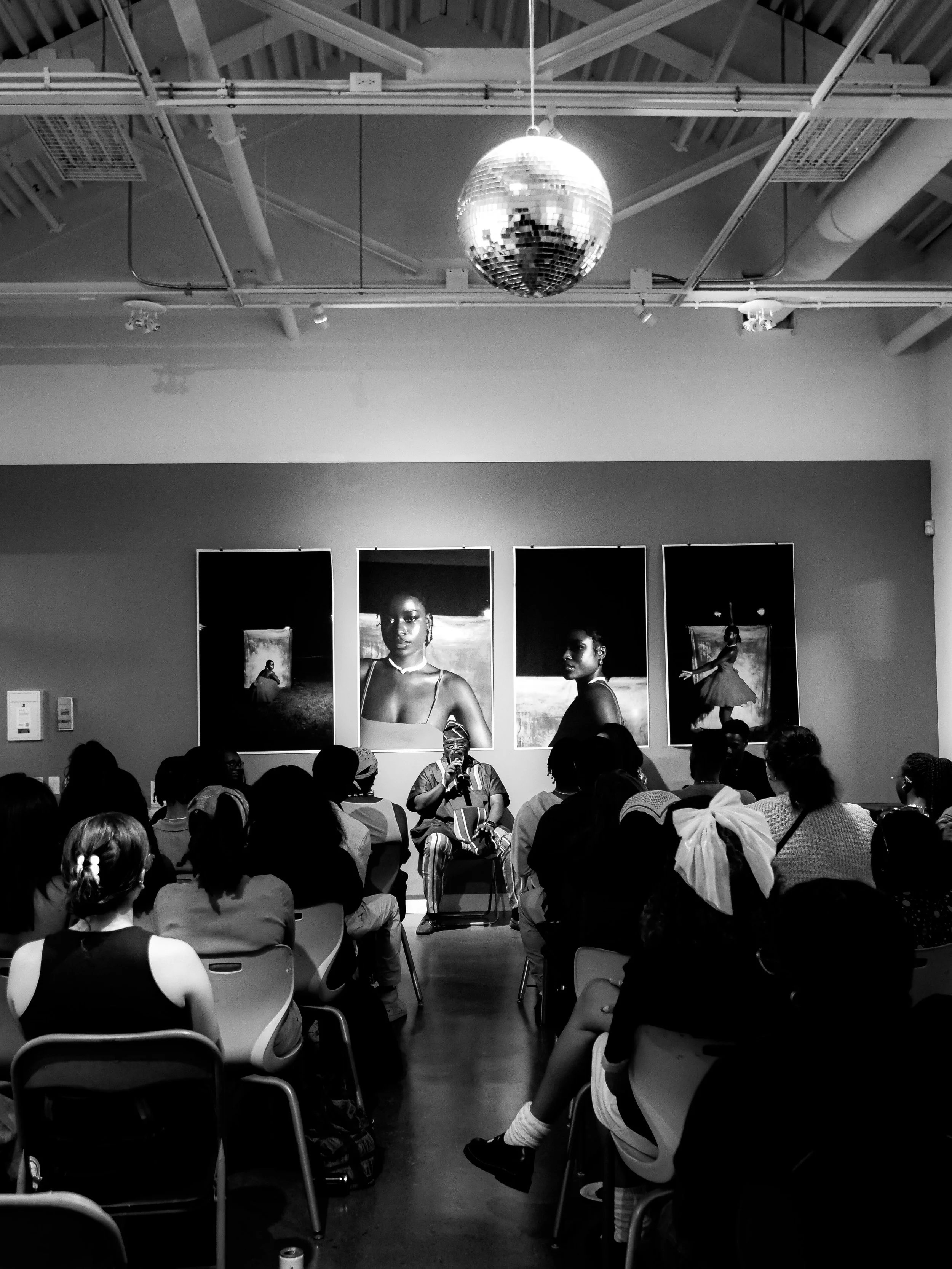 A speaker is seated in front of an audience in a gallery, with four large photographs of women displayed behind them. The gallery has a high ceiling with exposed piping and a hanging disco ball.