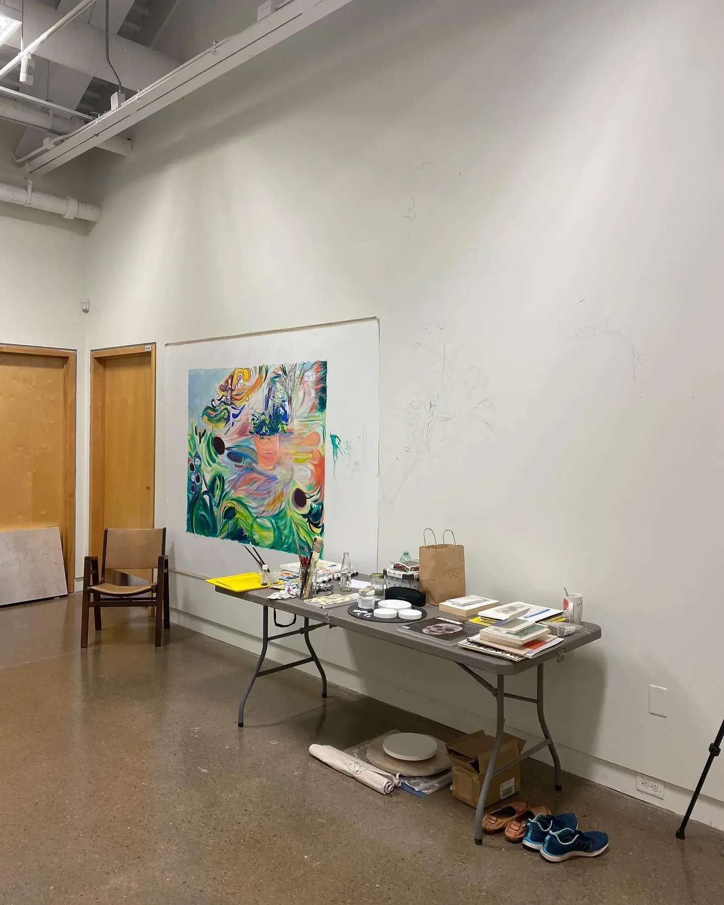 Art studio with a table of painting supplies, a colorful abstract painting on the wall, and a few chairs and shoes on the floor.