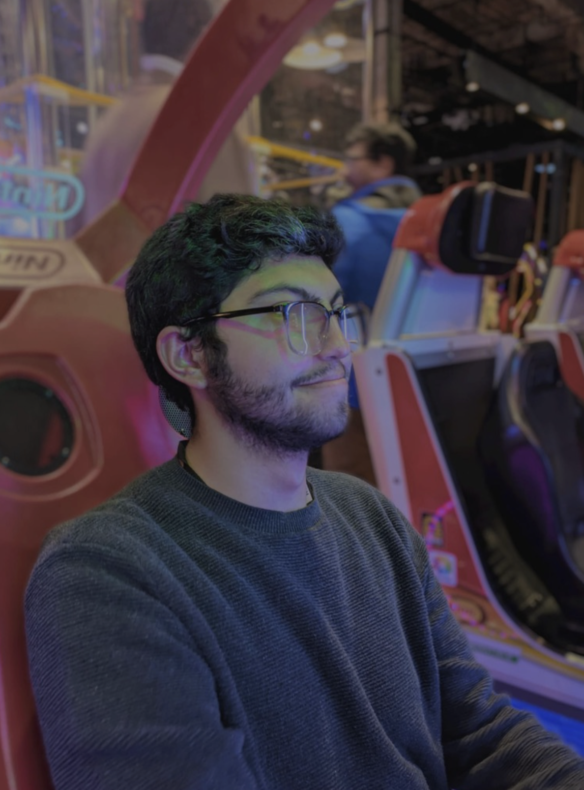 A man with dark curly hair and glasses, wearing a gray sweater, sitting at an arcade with colorful gaming machines around him, smiling with eyes closed.