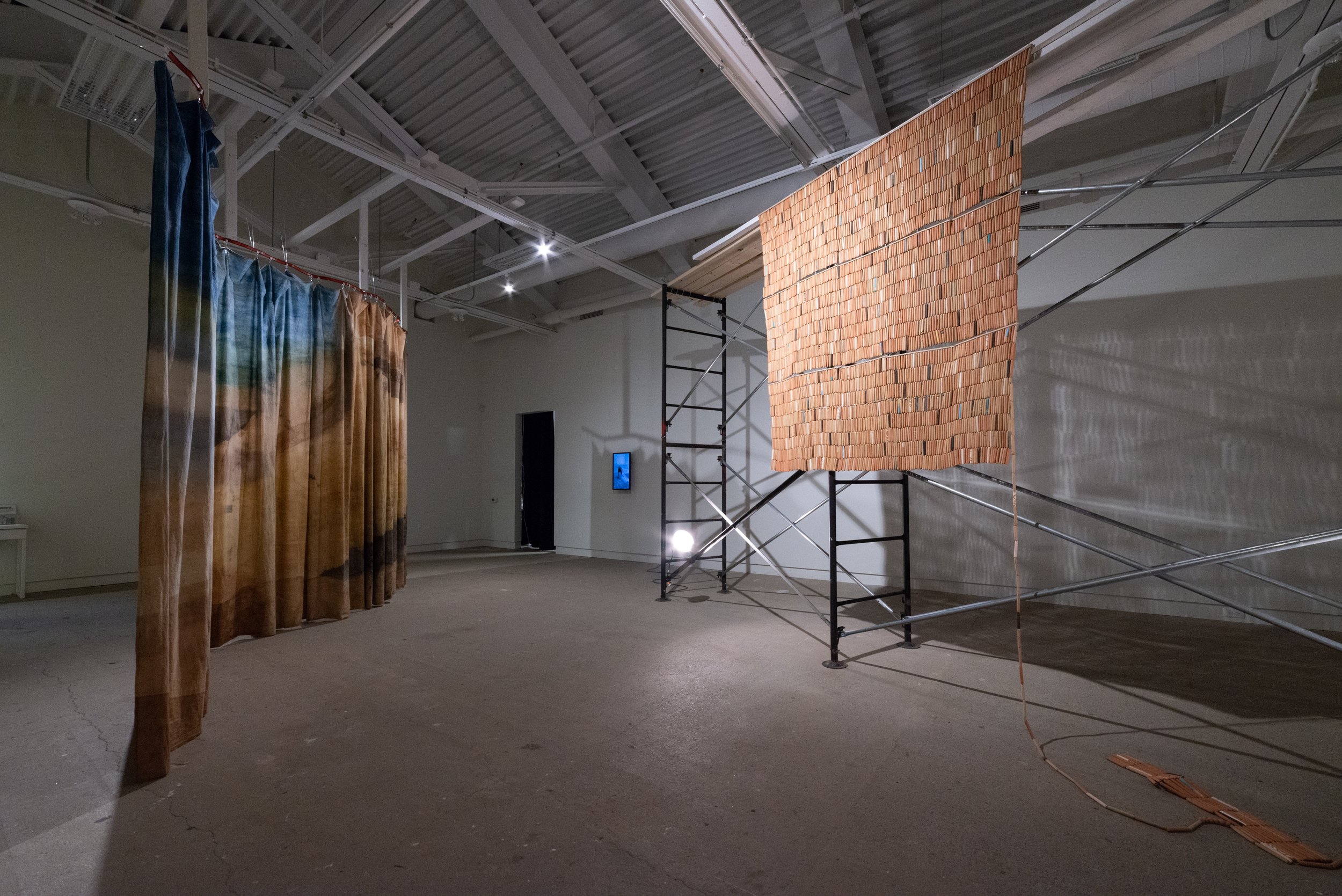 An art installation featuring a curved, multicolored curtain on the left, a large wooden mosaic panel on the right, and a smaller mosaic piece on the floor. The installation is set in a spacious gallery with high ceilings and exposed rafters.