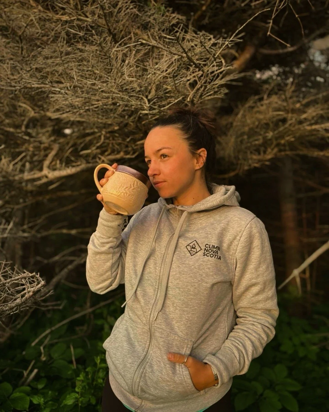 A woman wearing a gray hoodie with 'Climb Nova Scotia' logo holding a mug and standing outdoors near a large tree with bare branches.