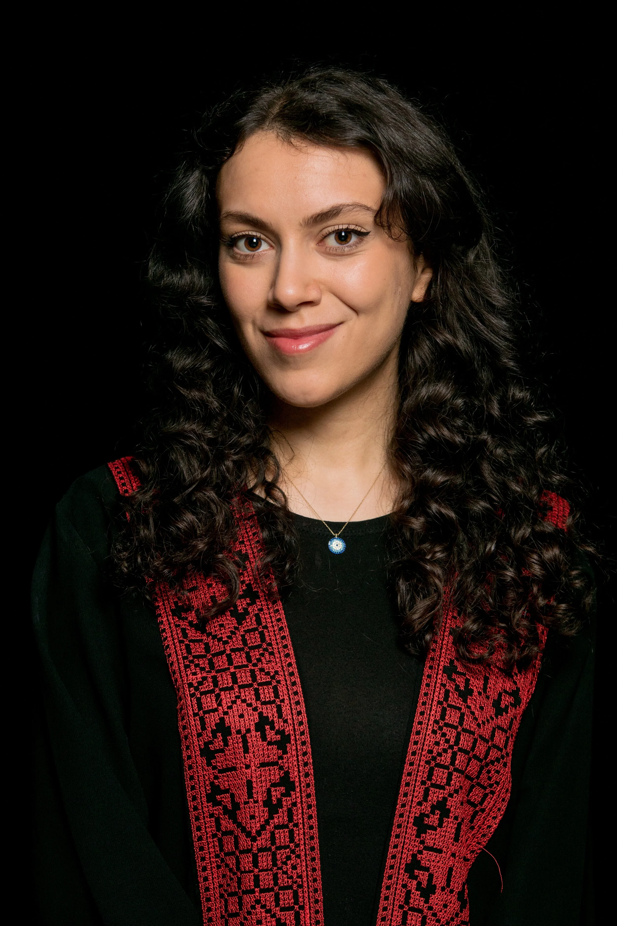 Portrait of a young woman with dark curly hair, wearing a black top with red embroidered accents and a blue and white religious pendant necklace, smiling against a black background.
