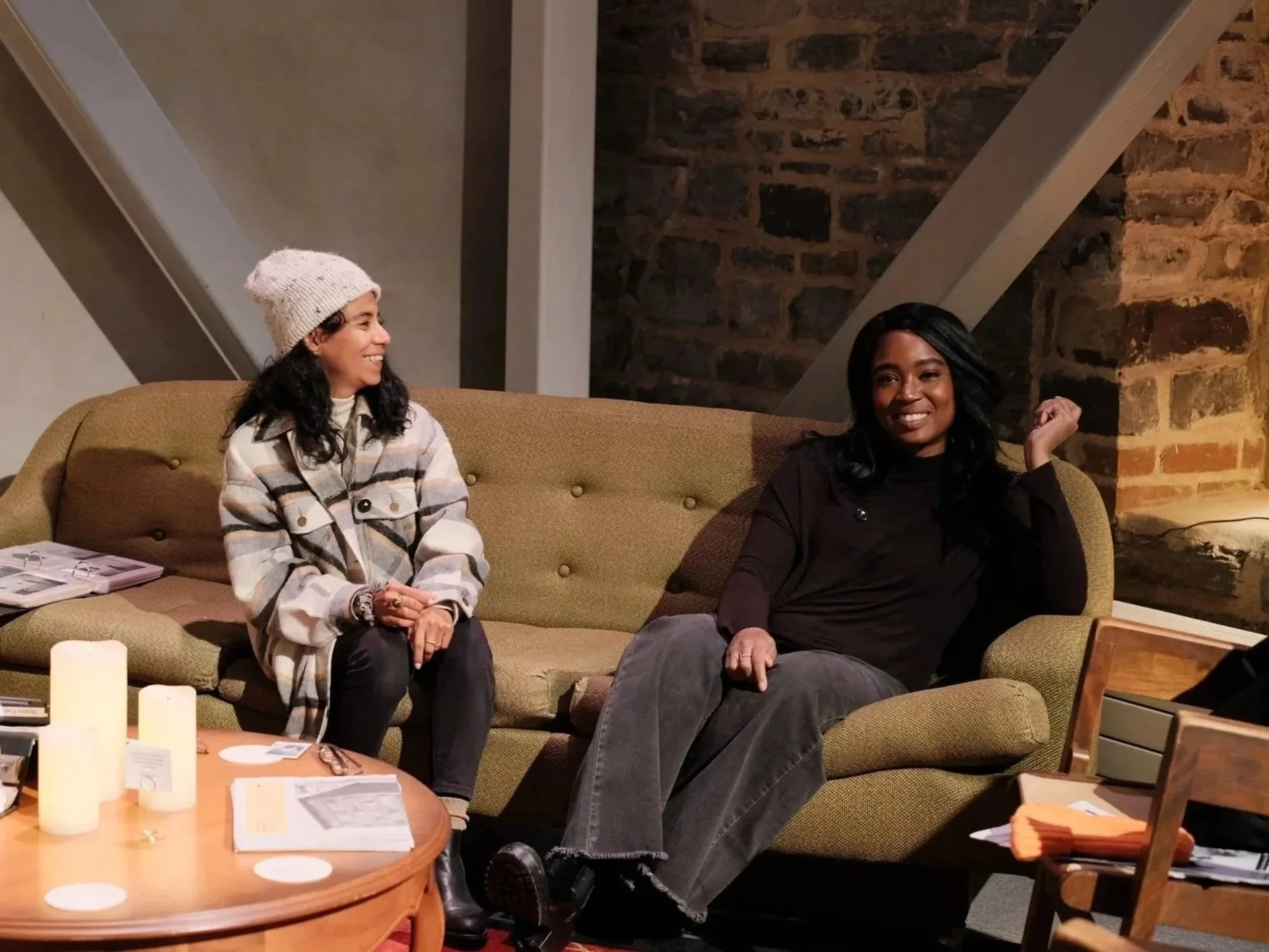 Two women sitting on a tan couch in a cozy room, one wearing a plaid jacket and a knit hat, the other in a black top. They are smiling and engaging in conversation. There are candles, papers, and other items on the coffee table in front of them.