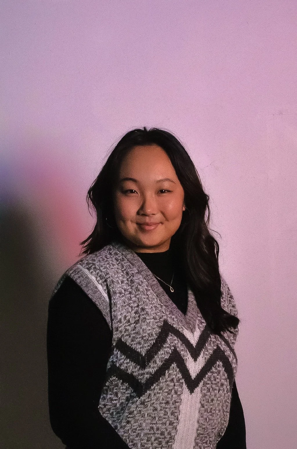 Portrait of a young woman with medium-length black hair, smiling slightly, wearing a black and gray patterned sweater over a black top, standing against a plain light pink background.
